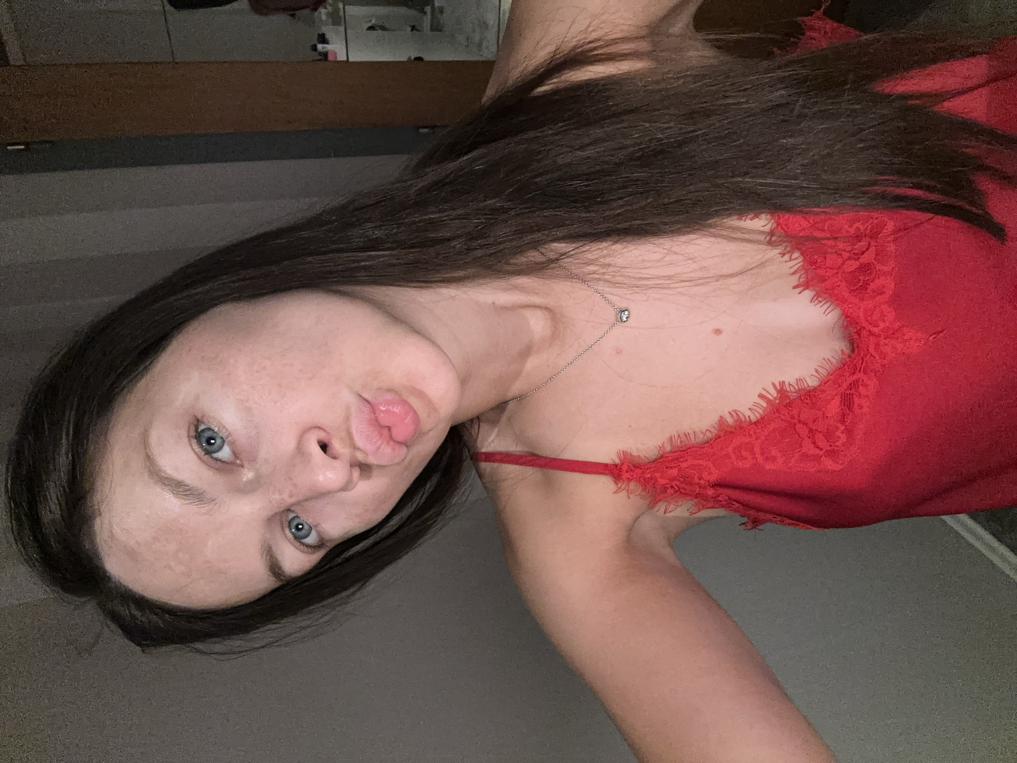 A woman takes a close-up, tilted selfie indoors, likely in Fredenbeck, Germany. She has long, dark brown hair, light blue eyes, and a fair complexion with visible freckles. Her lips are pursed in a "duck face" expression as she looks directly at the camera. She is wearing a bright red top, possibly a camisole or sleepwear, featuring intricate lace detailing along the neckline. A delicate silver necklace with a small, circular pendant adorns her neck. The background is dark and indistinct, with hints of a wooden surface and dark walls, suggesting an intimate, indoor setting, possibly a bedroom. The lighting is dim, indicating the photograph was taken in the evening or at night. The overall mood is playful and informal. No text is visible in the image.