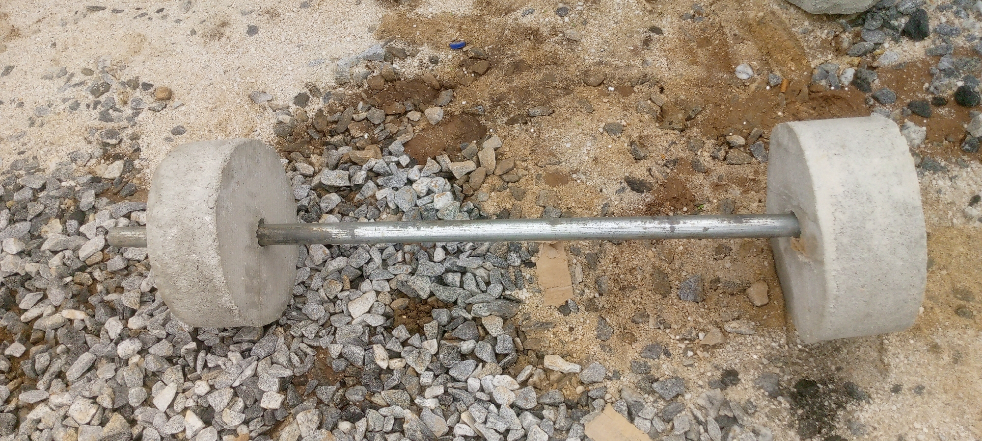The image shows a DIY dumbbell on the ground. The dumbbell consists of a metal bar with two concrete discs at either end, serving as weights. The dumbbell is lying on a surface composed of sand, gravel, and some dirt. The setting appears to be an outdoor environment, possibly a construction site or a backyard in Indo, Nigeria, given the materials and the rough terrain. There are no people visible in the image, and no actions or interactions are depicted. The lighting suggests it is daytime.