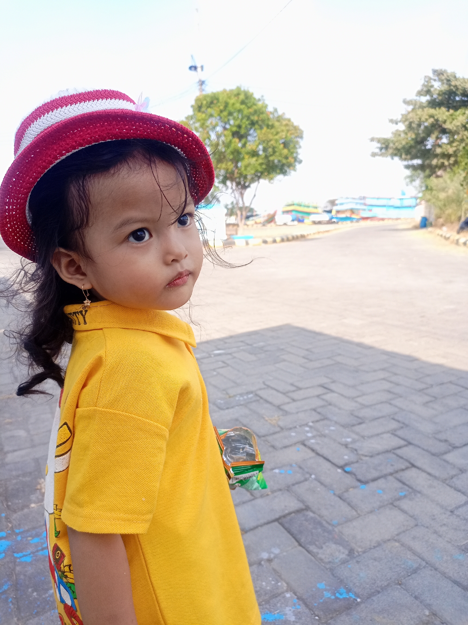 In Juwana, Indonesia, a young girl, approximately four years old, stands on a paved pathway. She wears a red and white striped hat and a bright yellow polo shirt. Her dark hair is styled in loose curls, and a small gold earring adorns her left ear. She holds a small plastic container of what appears to be food or candy. The background shows a paved road with a few trees and buildings in the distance. The sky is clear and bright, suggesting a sunny day.