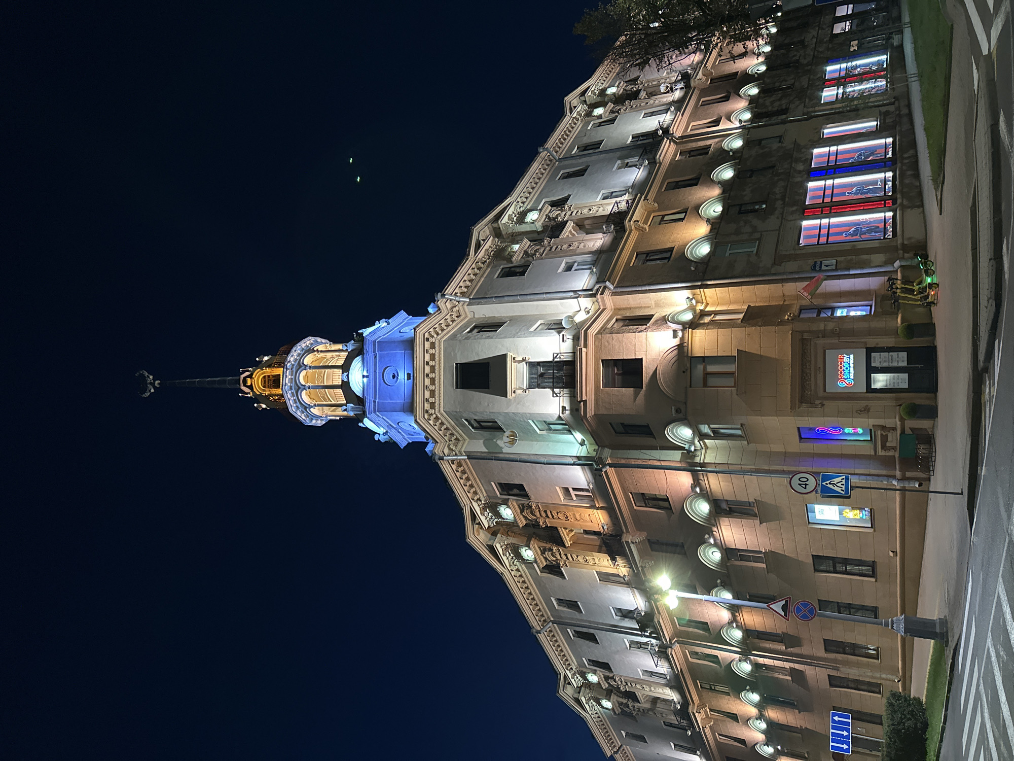 This is a low-angle, night-time exterior shot of a grand, corner building in Minsk, Belarus, likely a historical landmark. The building features a prominent spire topped with a crescent moon symbol, illuminated in yellow and blue light. The architecture is ornate, with classical details like columns and decorative moldings visible on the facade, all lit by warm-toned spotlights.
The building has multiple stories with numerous windows, some of which are displaying colorful advertisements or internal lighting. On the ground floor, there are storefronts, including one labeled "SOCCER SHOP.BY". Several traffic signs are visible on the street, including a speed limit sign showing "40" and pedestrian crossing signs. A crosswalk is prominent in the foreground. Two yellow electric scooters are parked on the sidewalk near the entrance to the soccer shop. The sky is dark blue, with faint green lights visible in the upper right quadrant, potentially aircraft or distant lighting. The overall impression is one of architectural grandeur illuminated against the night sky.