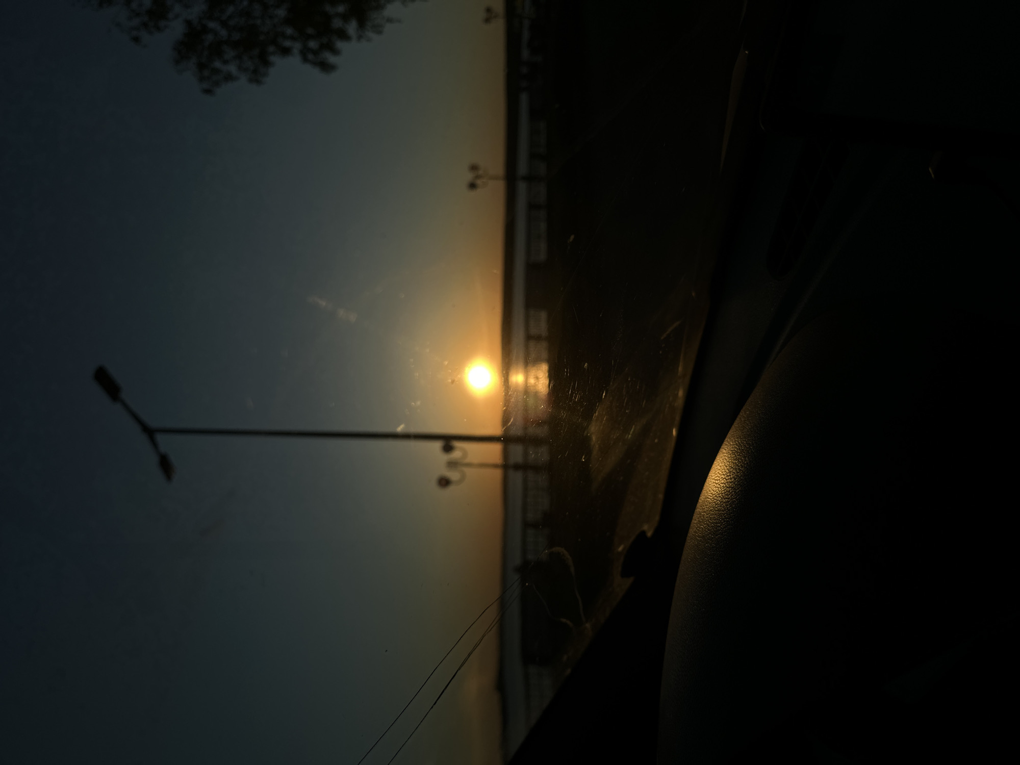 View from inside a vehicle at dusk with the setting sun over the city of Votkinsk, Russia. Streetlights are visible along the waterfront. The dashboard of the vehicle occupies the foreground.