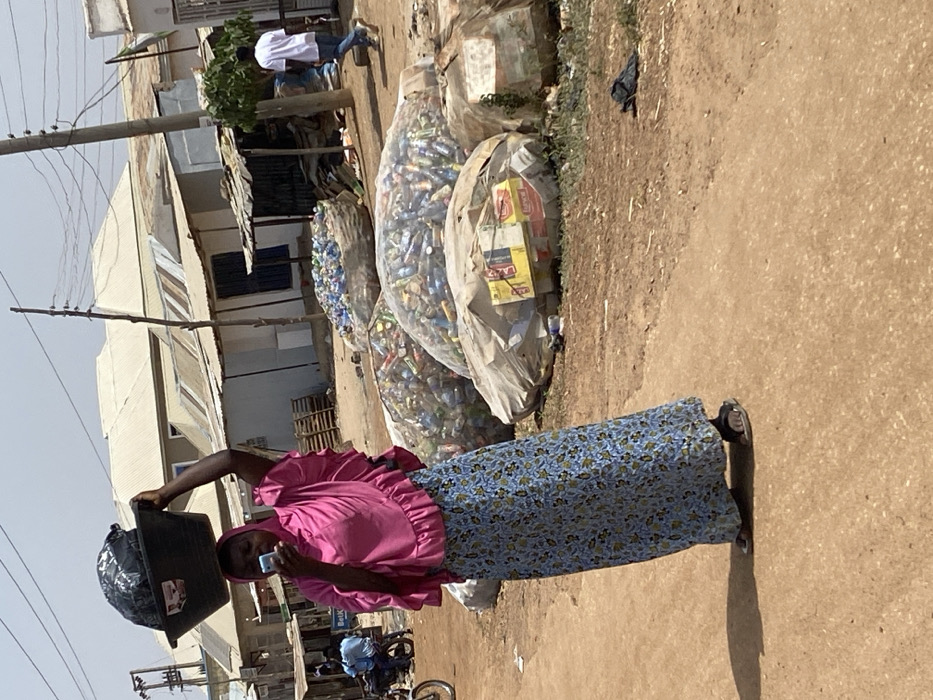This image depicts a sunlit, unpaved street scene in Baganakwo, Nigeria. The central figure is a woman positioned slightly left of center, walking towards the viewer's left. She wears a vibrant pink head covering that extends over her shoulders, a matching pink top with ruffled sleeves, and a long blue skirt with a floral pattern. She carries a black plastic basin containing dark material on her head, stabilized by her raised right hand. Simultaneously, she holds a light-colored smartphone in her left hand, viewing its screen.
To her right, a collection of large, translucent white bags are stacked on the dirt ground, filled with various recyclables, predominantly plastic bottles and aluminum cans. One bag contains cardboard boxes. Behind these bags, a rudimentary structure with a corrugated metal roof is visible. Further in the background, a man in a white t-shirt and blue jeans walks away from the viewer towards the right. Several single-story buildings with corrugated metal roofs and light-colored walls line the street in the distance.
To the left of the central figure, a utility pole with multiple overhead wires stands. Two individuals are seated on a motorcycle parked near a building in the mid-ground. A partially visible sign displaying "BetK" is on a building in the background. The ground is reddish-brown dirt, showing texture and dust, with distinct shadows indicating strong overhead sunlight.