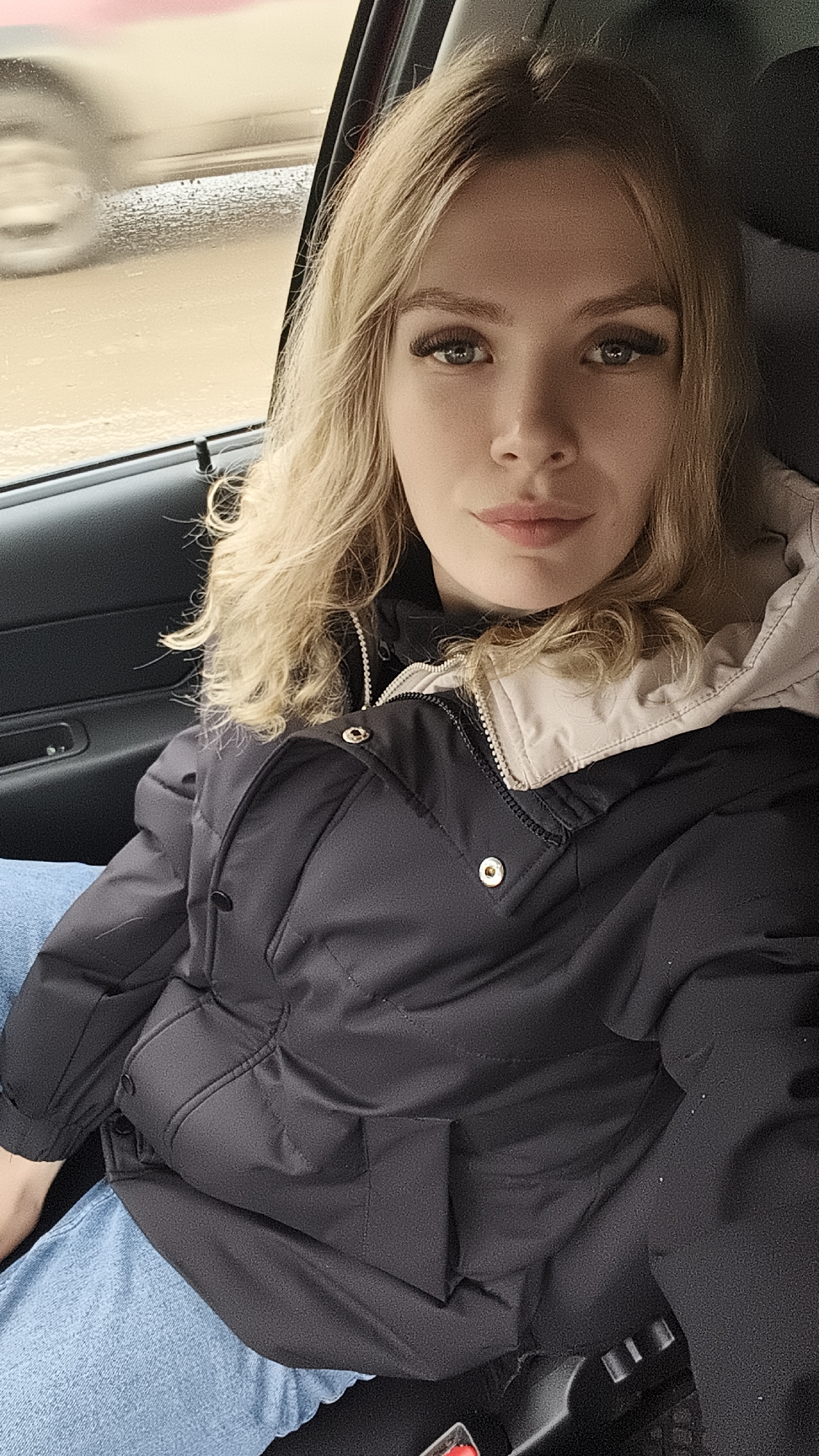 A young woman with blonde, wavy hair and light-colored eyes is depicted sitting in the passenger seat of a car. She has prominent eyelashes and a neutral expression, looking directly at the camera. Her hair is parted on the side and falls over her shoulders. She is wearing a black puffer jacket with a light beige or off-white hood, featuring gold-colored snaps or buttons and a visible zipper. Her light blue jeans are partially visible on her lap.
The setting is inside a car, with a dark-colored door panel visible to her left. Through the car window behind her, an outdoor scene is blurred, suggesting the car is moving or has recently been in motion. The background outside appears wet and muted, with greyish-brown tones, indicative of an overcast day or recent rain. A light-colored object, possibly another vehicle, is blurred in motion on the left side of the window. No visible text is present in the image. The scene suggests a casual moment, possibly a selfie taken while traveling in Izhevsk, Russia.