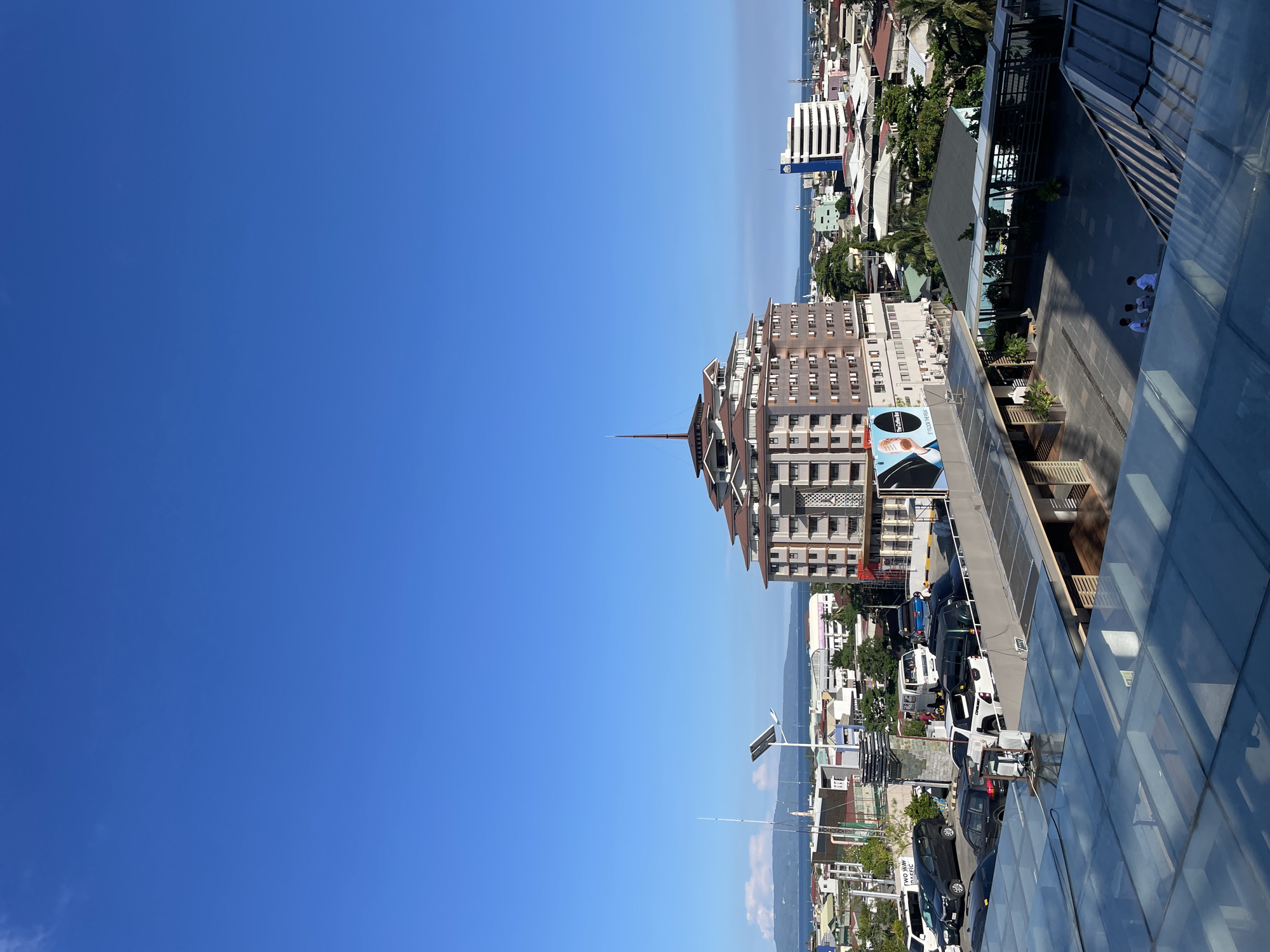 The image is an outdoor, high-angle shot of a cityscape with a clear blue sky. The scene is oriented sideways, likely a consequence of the original photo being taken in portrait mode and then rotated. In the foreground, a modern building with a glass exterior and dark framework occupies the right side of the frame. This building overlooks a courtyard area below, where three individuals, dressed in white tops and dark bottoms, are walking. Beyond this, the cityscape unfolds with numerous buildings of varying heights and architectural styles, interspersed with lush green trees. A distinctive, multi-layered building with a pointed spire is a prominent feature in the mid-ground. Further in the distance, a body of water, possibly the sea, is visible, meeting a hazy horizon. Cars are parked along a street running through the city. The time of day appears to be daytime, with bright sunlight casting shadows. The weather is clear and sunny. The location is identified as Davao City, Philippines. A sign with Asian characters and the text "No Coffee" is visible on a billboard.