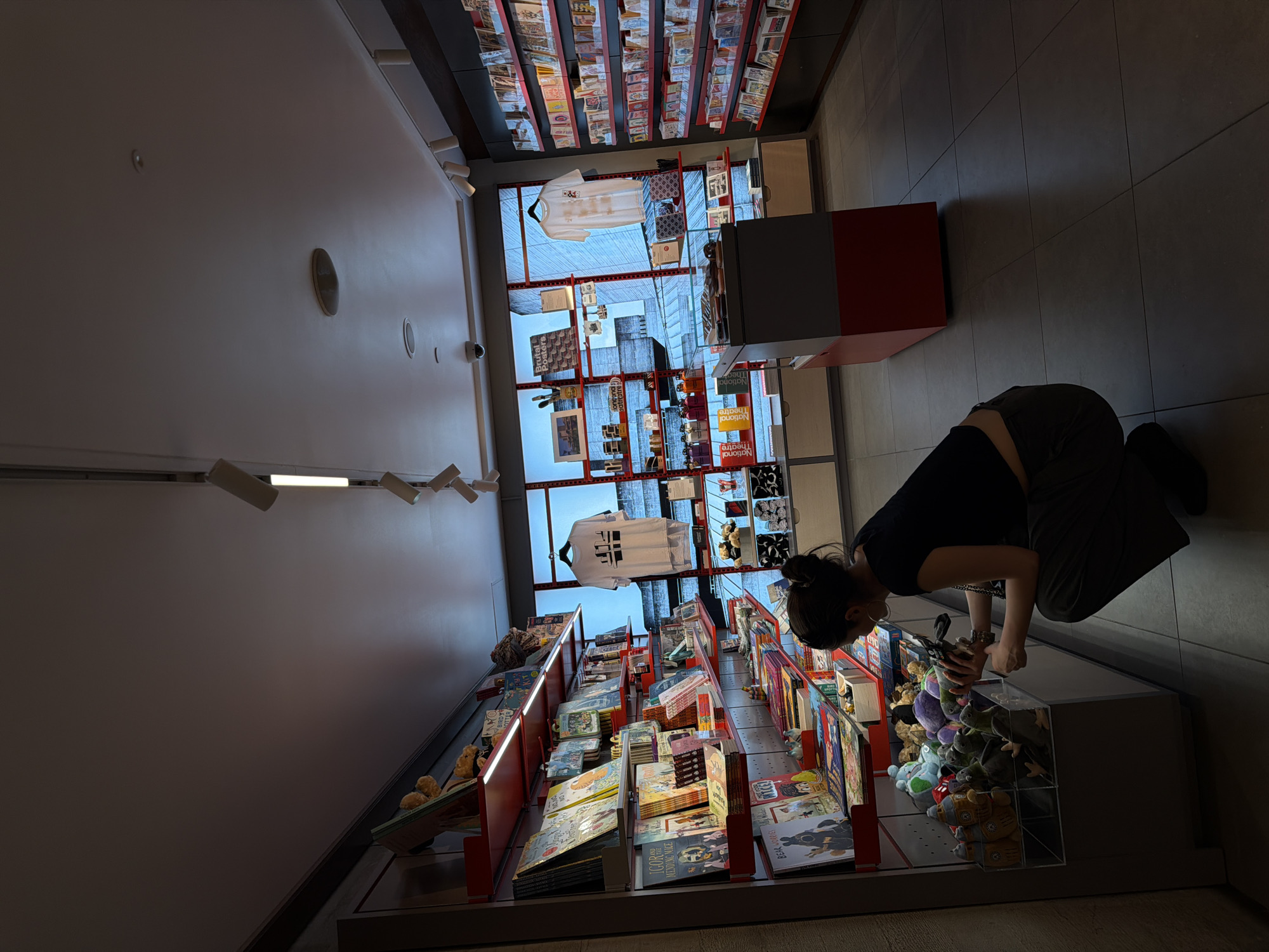 A woman in a black crop top and dark trousers bends to examine a display of plush toys in a shop. The shop appears to sell books and merchandise, evidenced by shelves filled with books and a t-shirt hanging in a window display. The location is likely the National Theatre Bookshop in London, United Kingdom, given the merchandise and the view through the window. The floor is tiled in dark gray squares. Overhead lights and ventilation fixtures are visible on the white ceiling.