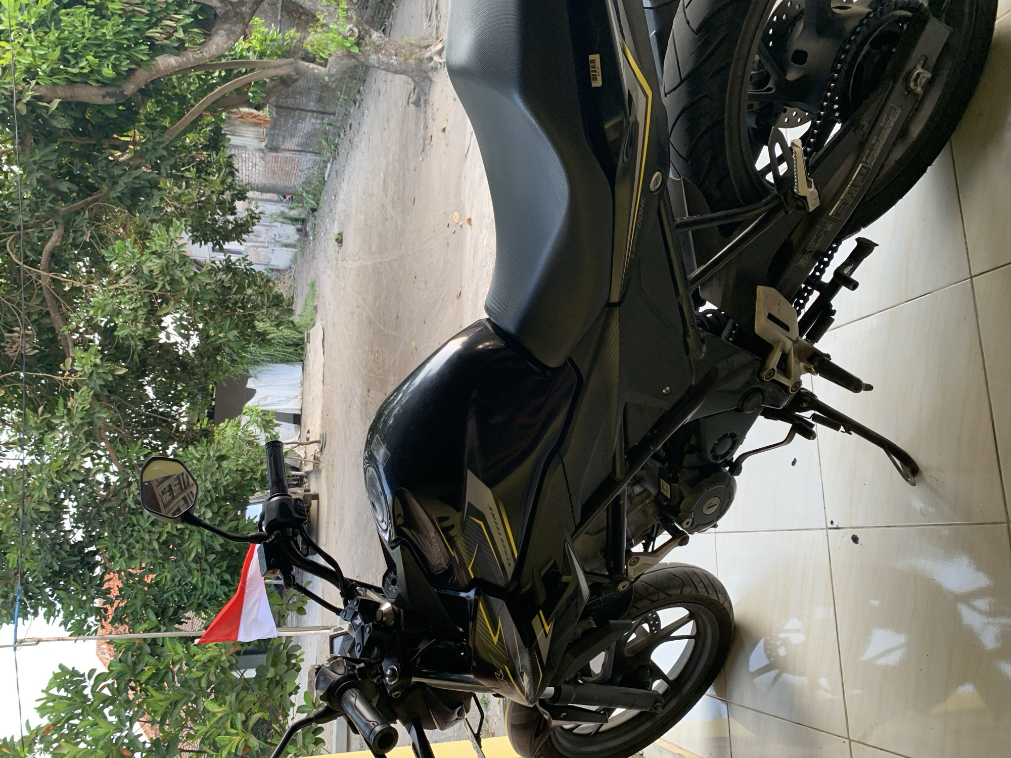 A black motorcycle is parked on a tiled patio in front of a dirt path. The motorcycle is facing right, with its front wheel slightly turned. A red and white flag is flying from a pole behind the motorcycle, to the left. The background shows trees, a brick wall, and a building with a white tarp. The scene appears to be in a residential area in Rejoso, Indonesia.