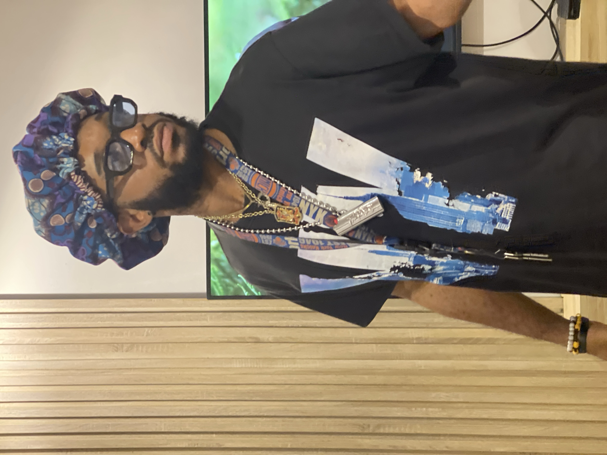 The image shows a person wearing a black t-shirt with a blue and white graphic print. They are also wearing a colorful satin bonnet and sunglasses. Around their neck, the person is wearing several necklaces, including a chunky silver chain and a gold chain with a pendant. A lanyard with "NYK" and other text is also visible. On their wrist, they are wearing bracelets, one with yellow and black beads. The person is standing in front of a wall with horizontal wooden paneling and a screen displaying a nature scene. The setting appears to be indoors, possibly an office or a presentation room. The lighting suggests it is daytime. The person's expression is not clearly discernible due to the sunglasses.