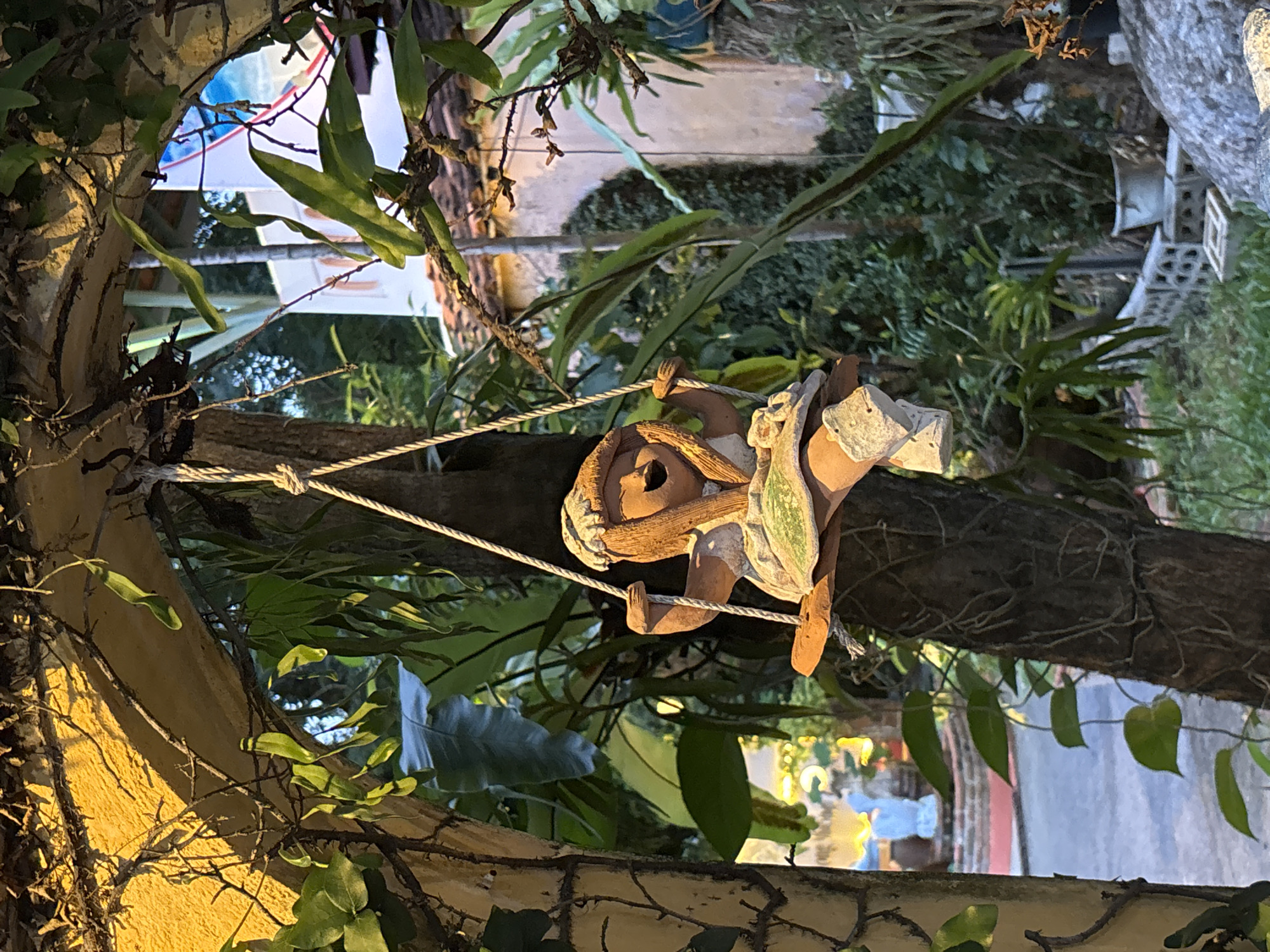 A ceramic doll is depicted on a swing, suspended from a tree. The doll, with a smiling expression and wearing a cap and dress, is positioned in the center of the frame. The swing is made of wood and tied with rope. The background reveals lush green foliage, suggesting a garden or outdoor setting. The scene is framed by an arched structure, possibly part of a building or a decorative entrance. The lighting indicates it may be daytime, with shadows cast by the surrounding vegetation. The doll's playful pose on the swing evokes a sense of childhood innocence and joy. The location is identified as Chalong, Thailand.