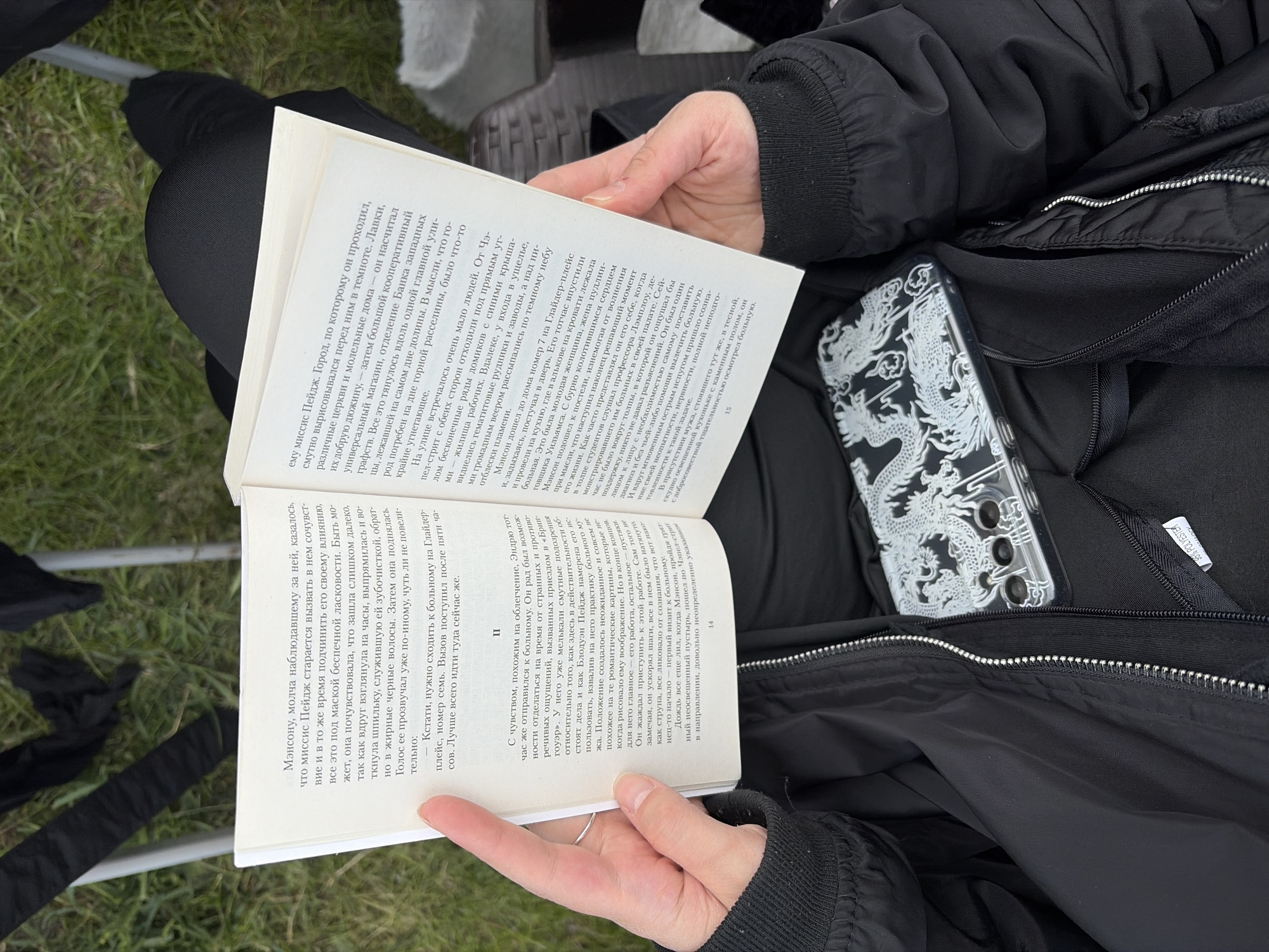 Here is a description of the image:
A person is seated, likely outdoors, reading a book. They are wearing a black jacket and black pants. The book is open, displaying text in Russian, and is positioned in front of them. A cell phone in a clear dragon patterned case is visible. The person is holding the book with both hands. The environment appears to be a mix of natural and constructed elements.