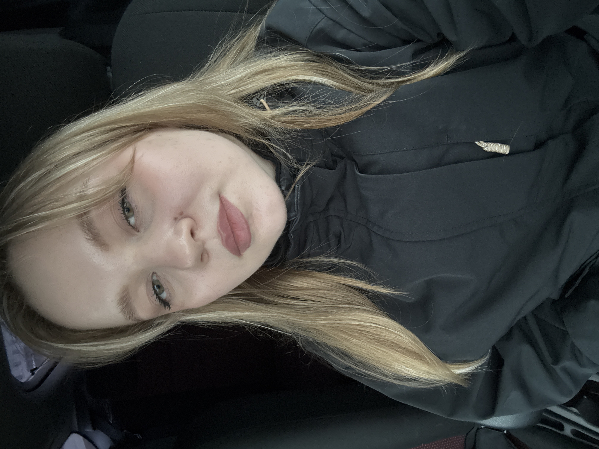 A young woman with long, straight blonde hair and light green eyes is the primary subject of this close-up, rotated selfie. She is looking directly at the camera with a soft, slight smile. She wears a dark, possibly black, casual jacket or coat, which covers most of her upper body. Her makeup is light, featuring defined eyebrows, subtle eyeliner on her upper lids, and a natural-toned lipstick.
The setting appears to be inside a vehicle, suggested by the dark, textured upholstery visible behind her head and shoulders, which resembles car seats. The image captures her from approximately the chest up. Her blonde hair cascades over her shoulders and jacket. A small, light-colored, coiled object, possibly a drawstring or a decorative element, is visible on her jacket near her right shoulder.
The lighting is soft and diffused, indicating it might be daytime with overcast skies, or an interior setting with indirect light. No specific time of day or weather can be definitively determined. The photograph exudes a calm and relaxed mood. There is no visible text in the image. Given the provided context, the location of this photo is Chaikovsky, Russia.