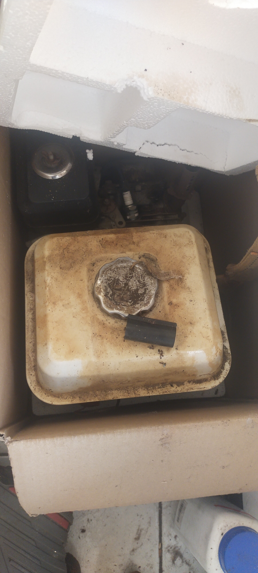 The image shows a close-up, overhead view of a small, weathered engine, likely from a generator or lawnmower, packed within a cardboard box. The engine's fuel tank, a pale yellow color, is prominently displayed and shows signs of dirt and grime. A silver, circular fuel cap is centered on the tank. To the right of the cap, a short, black rubber hose fragment rests on the tank's surface. Near the top of the image, the engine's spark plug, with white ceramic insulation and a metal tip, is visible, along with other engine components. The box itself appears to be made of plain corrugated cardboard. The background is partially obscured by white, fragmented styrofoam packaging material. The scene is consistent with an item being stored or transported in a shipping container or storage unit, possibly in a workshop or garage environment. No specific location in Pasirhuni, Indonesia, can be identified from the visual evidence provided.