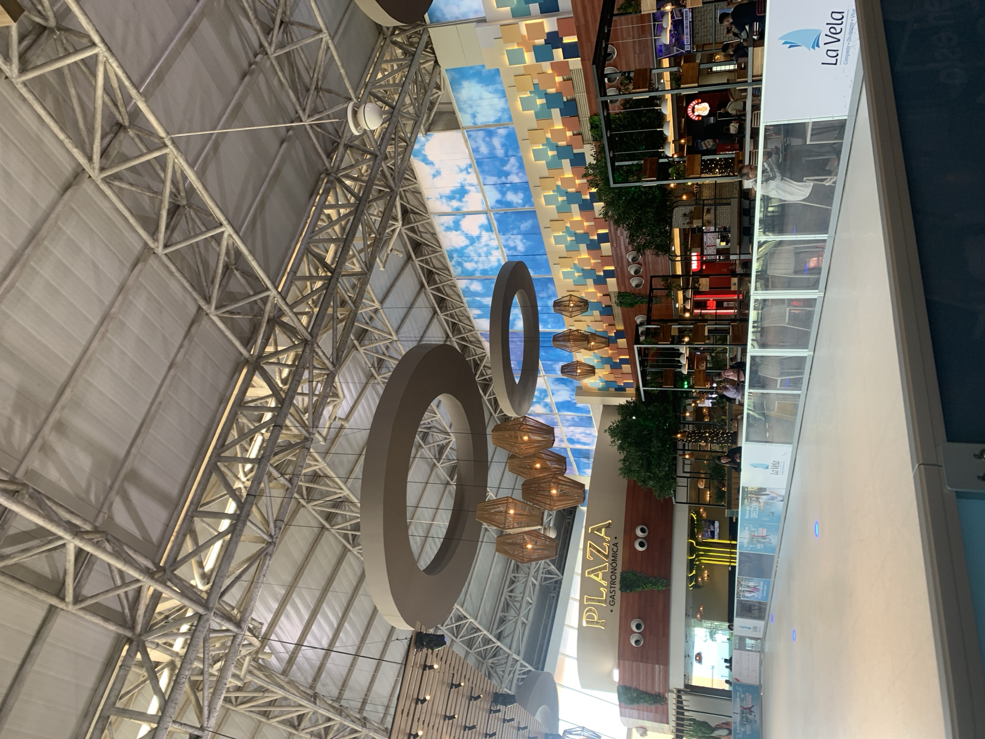 The image shows an interior view of a shopping mall. The mall has a high, vaulted ceiling with exposed metal beams. Circular light fixtures hang from the ceiling, alongside modern light fixtures. Sections of the ceiling are decorated with blue and white square patterns and a faux sky mural. On the right side of the frame, there is a multi-level section with restaurants and shops; "La Vela" is visible on one of the signs. The word "PLAZA" is also visible on a wall decorated with plants and two cartoonish eyes. People can be seen walking and sitting in the restaurants. The image is presumably taken in or near Porlamar, Venezuela, based on the "La Vela" branding, a known establishment in the area.