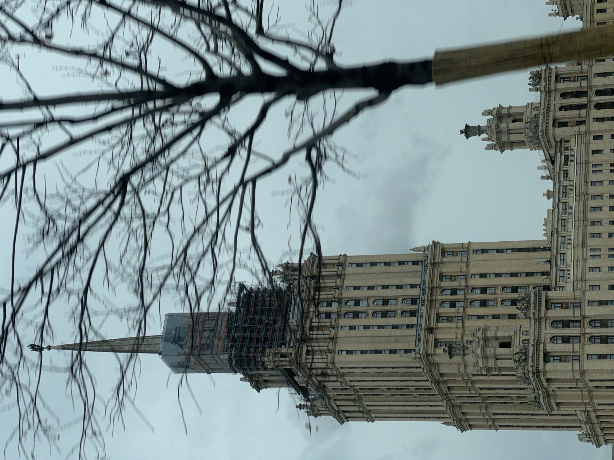 The image depicts a tall, ornate building in Moscow, Russia, likely one of the "Seven Sisters" Stalinist skyscrapers. Scaffolding surrounds the building's top portion, suggesting ongoing construction or renovation. Leafless tree branches fill the foreground, partially obscuring the view of the building. The sky is overcast, creating a subdued and somber atmosphere.