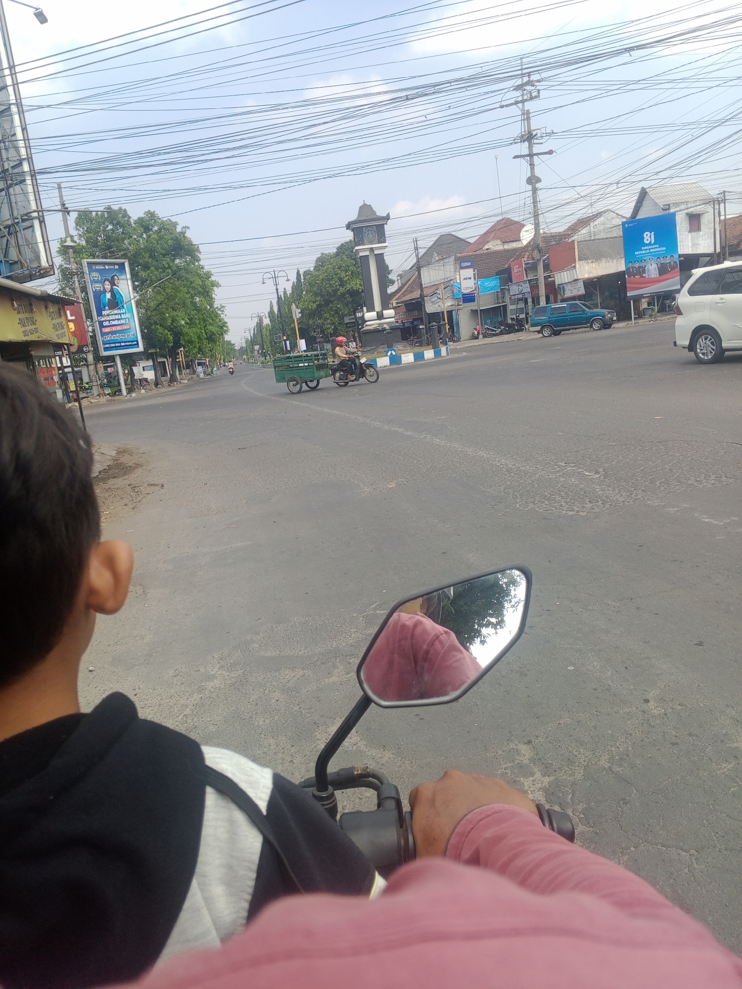 The image captures a street scene in Kadipaten, Indonesia, from the perspective of someone riding a motorcycle. In the foreground, the back of the rider's head and shoulder are visible, along with the handlebars and a pink sleeve. A motorcycle's side mirror reflects a person wearing a pink shirt and trees. The street is paved and appears to be a main road with traffic. Several vehicles are present, including a green trailer being pulled by a motorcycle, a blue SUV, and a white car. Buildings line the sides of the road, with a prominent clock tower in the distance. Billboards and signs are visible, including one with "81 DIRGAHAYU REPUBLIK INDONESIA". Power lines crisscross the sky. The environment suggests a typical day in an Indonesian city.