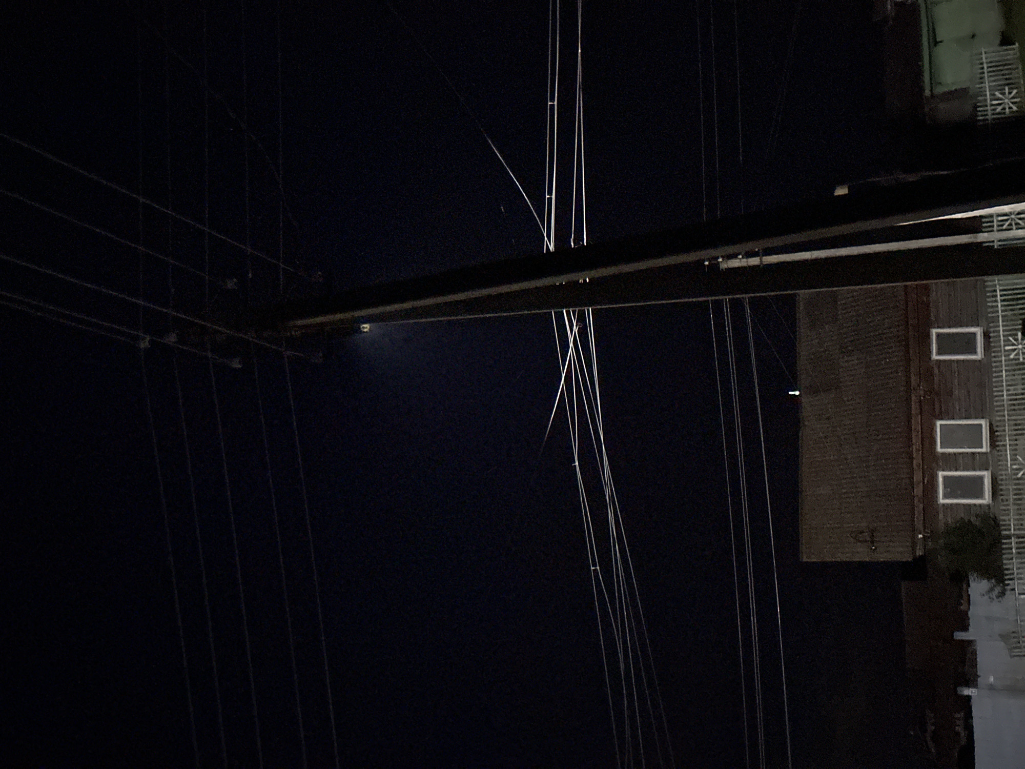 The image is a night time, low-light outdoor shot of a rural scene. A wooden utility pole with power lines stretching across the frame is the dominant object, positioned slightly off-center to the right. The pole itself is dark and textured, with what appear to be insulators attached to it. Several thin, light-colored wires are visible, crisscrossing against the dark blue-black night sky. In the lower half of the image, a dark, weathered wooden building with a corrugated roof is visible. Two rectangular windows with white frames are present on the building's facade. To the left of the building, a portion of a white picket fence is visible. A small, dark green building with a white door or garage is partially visible on the far right. The overall impression is one of a quiet, possibly isolated, rural area at night, illuminated only by ambient light and possibly distant artificial sources. The scene is devoid of any visible people or animals.