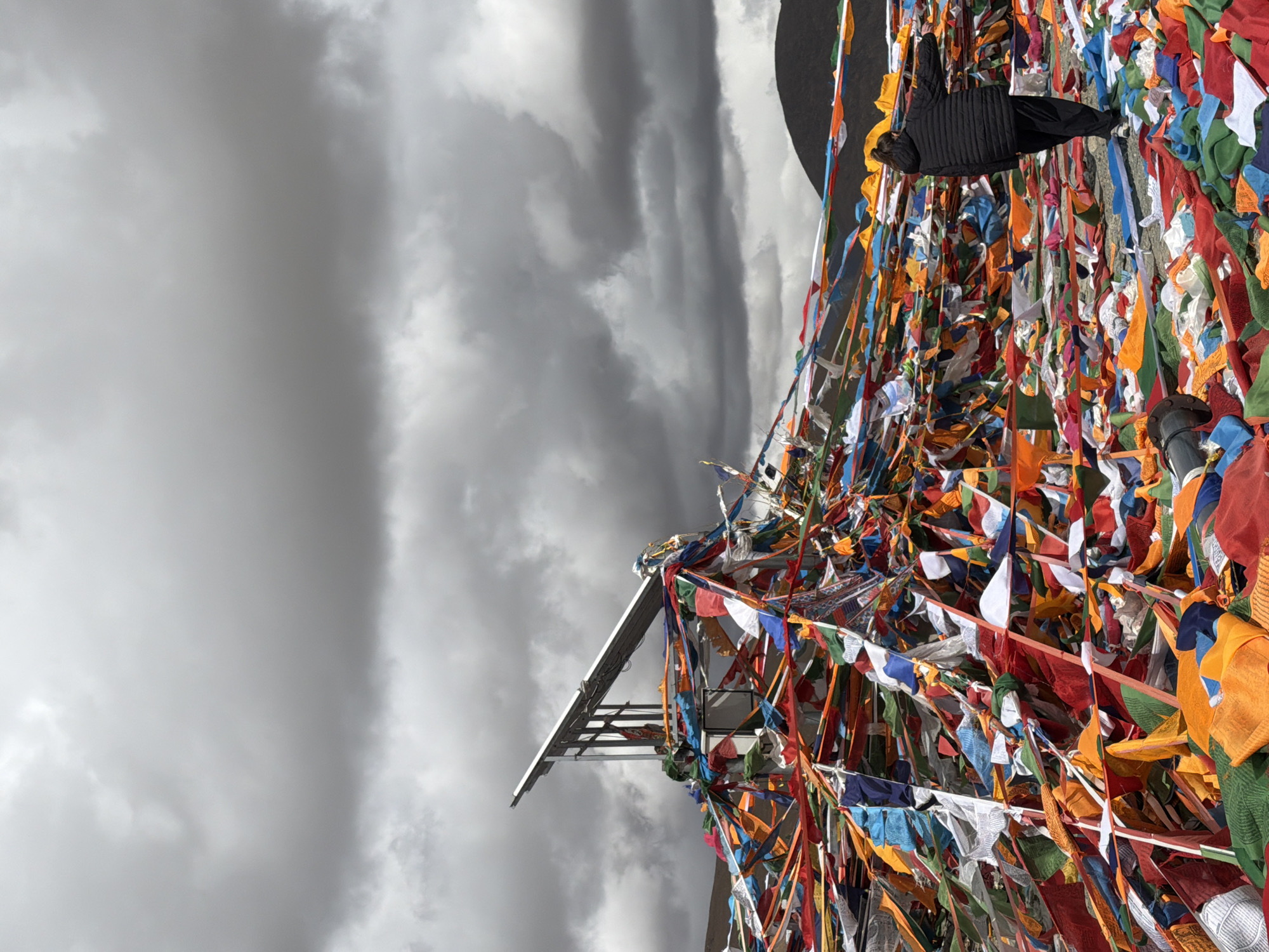The image shows a scene in Hor town, China. Prayer flags of multiple colors are strung on metal poles and structures, creating a dense display. A black jacket is affixed to the structure amidst the flags. The background is a cloudy, overcast sky.