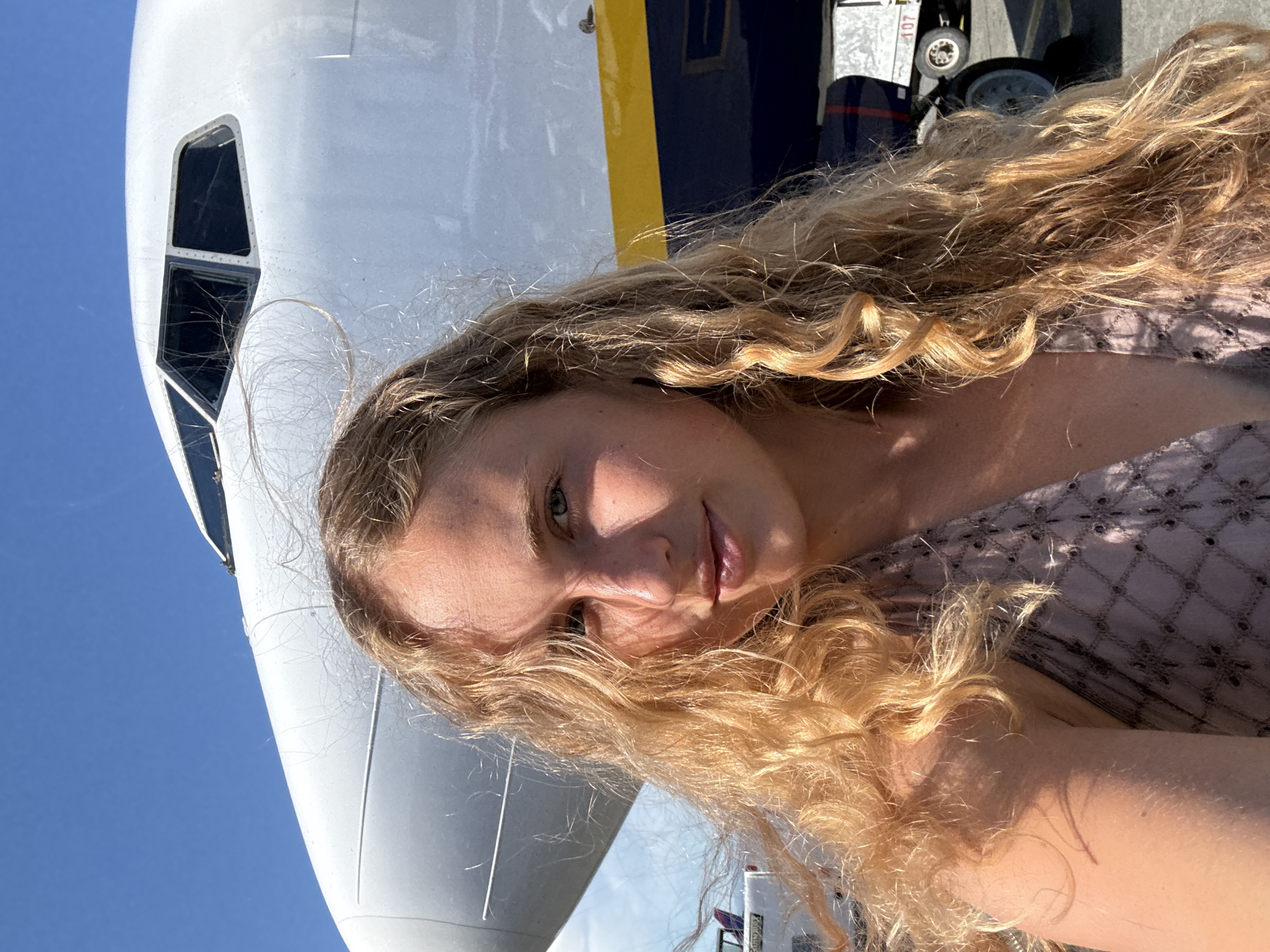 The image shows a close-up, selfie-style shot of a young woman with long, blonde, curly hair. She is positioned in front of the nose of a large aircraft, with the blue sky visible behind it. The woman's face is turned towards the camera, and she has a slight smile. Sunlight illuminates her face, creating highlights and shadows, suggesting it is daytime.
The aircraft appears to be a passenger jet, with its distinctive white fuselage and windows visible. A portion of the yellow and dark blue livery of the aircraft is also visible, hinting at an airline's branding. In the background, a wheel and part of the landing gear of the aircraft are discernible, along with some equipment or structures on the tarmac. The overall setting appears to be an airport or an area where aircraft are parked. The image captures a casual moment, possibly before or after a flight, with the woman seemingly enjoying the sunshine. The presence of the aircraft and the open sky suggest travel or a departure. No visible text is clearly legible within the frame.