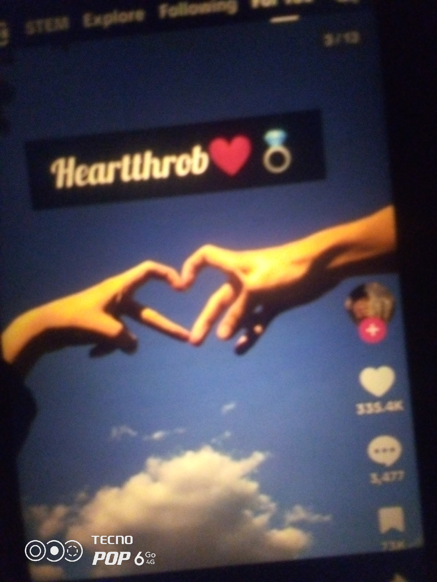 The image displays a close-up of a mobile phone screen, with a dark, possibly nighttime, environment surrounding it. The phone displays a social media interface, likely a video or image sharing platform. Prominently featured is the text "Heartthrob" in a stylized font, accompanied by a red heart icon and a diamond ring icon. Below this, two hands are visible, forming a heart shape against a blurred background of sky and clouds. On the right side of the screen, icons for likes, comments, and shares are present, with numerical counts indicating engagement. The lower portion of the screen shows a watermark for "TECNO POP 6 4G". The overall scene suggests a personal moment captured on a phone, with the user likely interacting with or viewing content related to relationships or love. There is no information within the image to indicate the location as Howayi, Nigeria.