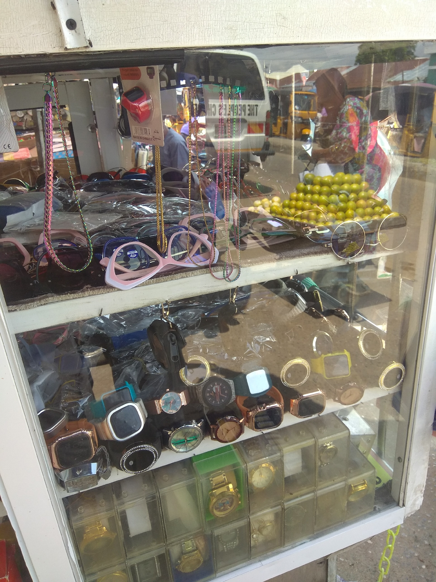 In Jalingo, Nigeria, a street vendor's stall displays an assortment of accessories behind glass. The stall is organized with multiple shelves showcasing various items. On the top shelf, a red digital watch is prominently displayed. Below this, a collection of sunglasses in different styles and colors are arranged. Further down, an array of wristwatches, both analog and digital, are laid out, many with decorative elements. Some watches are presented in individual clear plastic cases, while others are displayed directly on the shelf. A pile of small green fruits, possibly limes or small oranges, is also visible on the top shelf, adding a touch of vibrant color to the scene. Reflections on the glass show a busy street with vehicles, including a white van and yellow tricycles, suggesting a commercial area. A person wearing a colorful headscarf and patterned top is partially visible in the reflection, likely the vendor. The stall itself appears to be an outdoor setup, typical of informal markets in Nigeria.