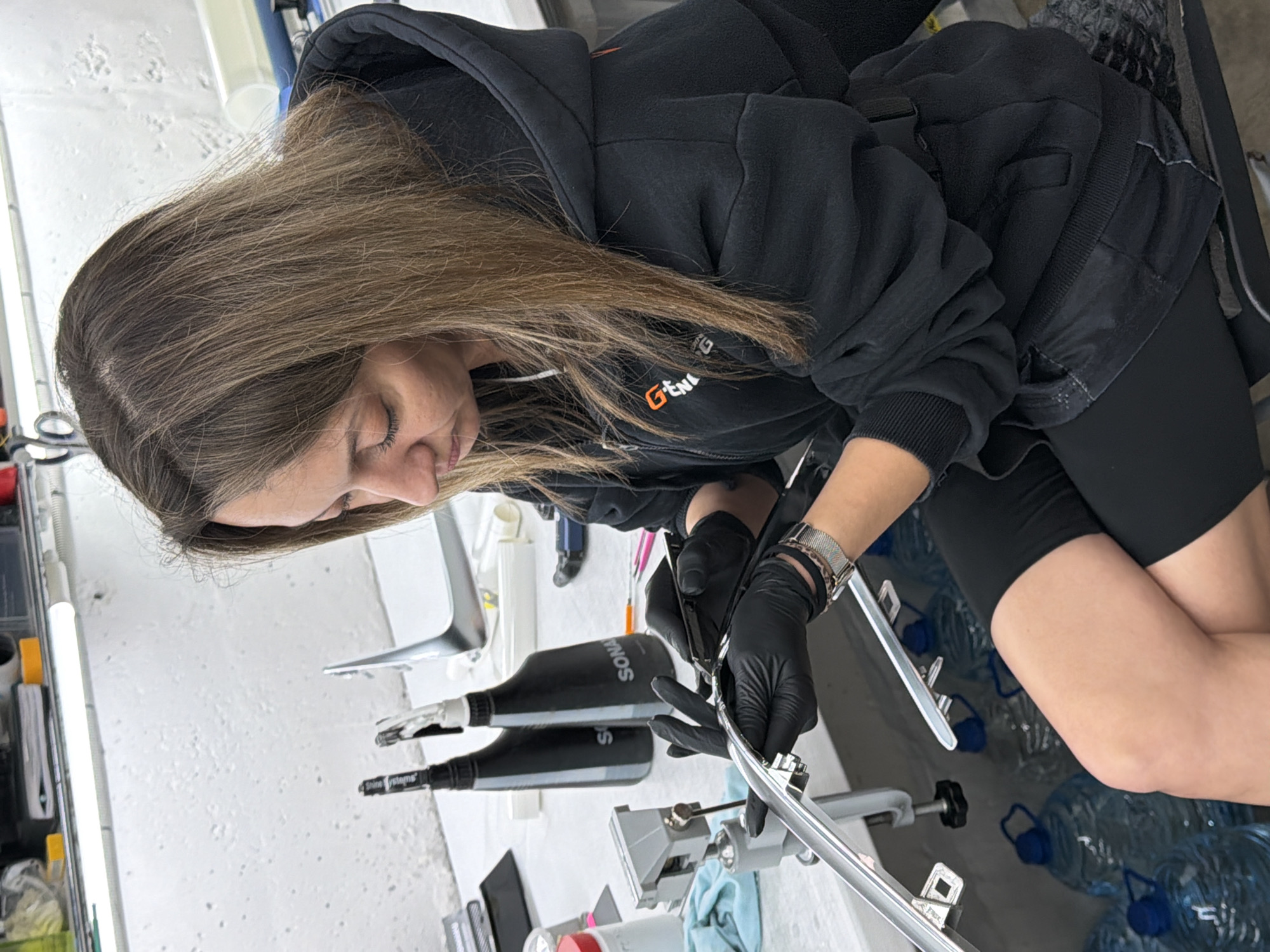 An adult female, characterized by light brown hair, is seated on a dark stool within what appears to be a workshop or detailing facility. She wears a dark grey/black hooded sweatshirt with an orange "G-Energy" logo on the left chest, black athletic shorts, and black disposable gloves. A silver-toned watch with a patterned band is visible on her left wrist. The woman is intently focused on a silver, reflective automotive trim component, which is secured by a grey clamp/vise on a silver stand attached to a white workbench. She is utilizing a small, black-handled tool in her right hand to work on the component. The workbench also holds two black spray bottles prominently labeled "SONAX," alongside scissors, red and pink-handled detailing tools, a red-capped container, a light blue rag, a black rectangular tray, and a roll of transparent film on a blue tube. The immediate background features a white, textured wall illuminated by an overhead fluorescent light fixture. Beneath the workbench, multiple clear plastic five-gallon water bottles with blue caps are stored. This scene is located in the city of Krasnoyarsk, Russia.