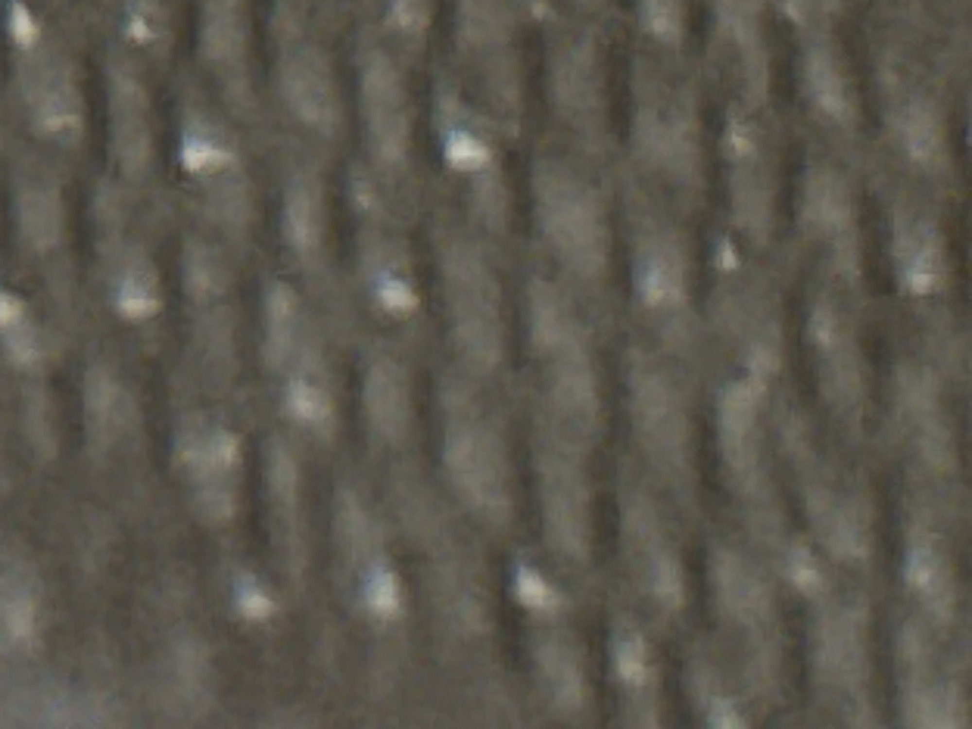 The image shows a close-up, macro shot of a textured gray fabric. The fabric has a raised pattern, with columns of raised lines separated by recessed channels. There are small, bright highlights scattered across the surface, suggesting a slightly reflective material or light catching on the texture. The texture is uniform and appears to be a woven or knitted material. No people, animals, or identifiable objects are visible. The background is entirely filled by this fabric texture. The image does not contain any visual information that would allow for identification of its location within the city of Navbahor, Uzbekistan.