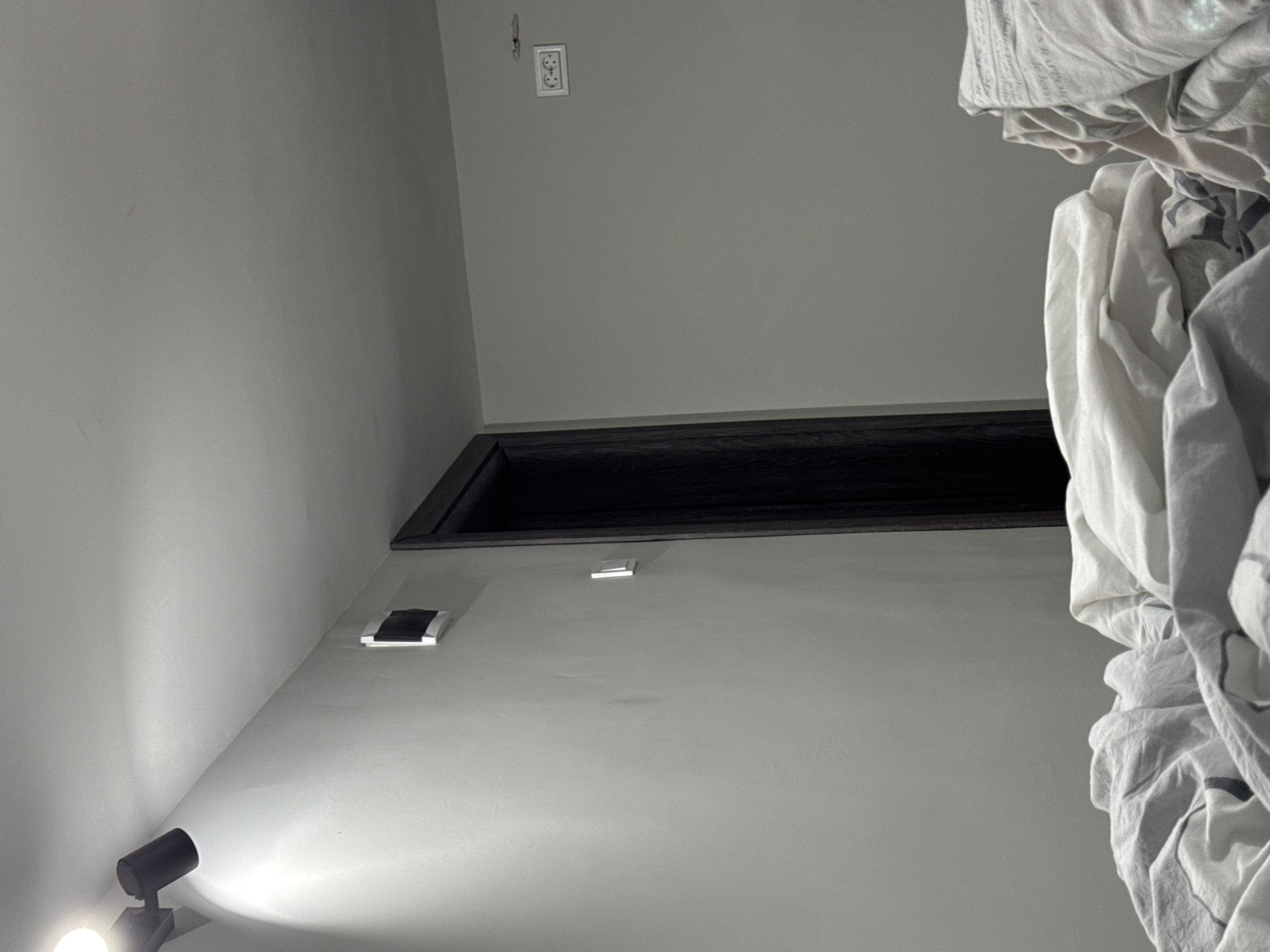 The image is an upward shot of an interior space, predominantly gray in color, potentially in Lovech, Bulgaria. The left side features a gray wall with a directional spotlight attached at the bottom corner, emitting a bright light. Above, on the same wall, are two small square light fixtures or sensors, and further up there is a black recessed area near the ceiling. In the upper part of the frame, there's an electrical outlet. The right side is dominated by a heap of white fabric, likely bedding. No people or interactions are visible.
