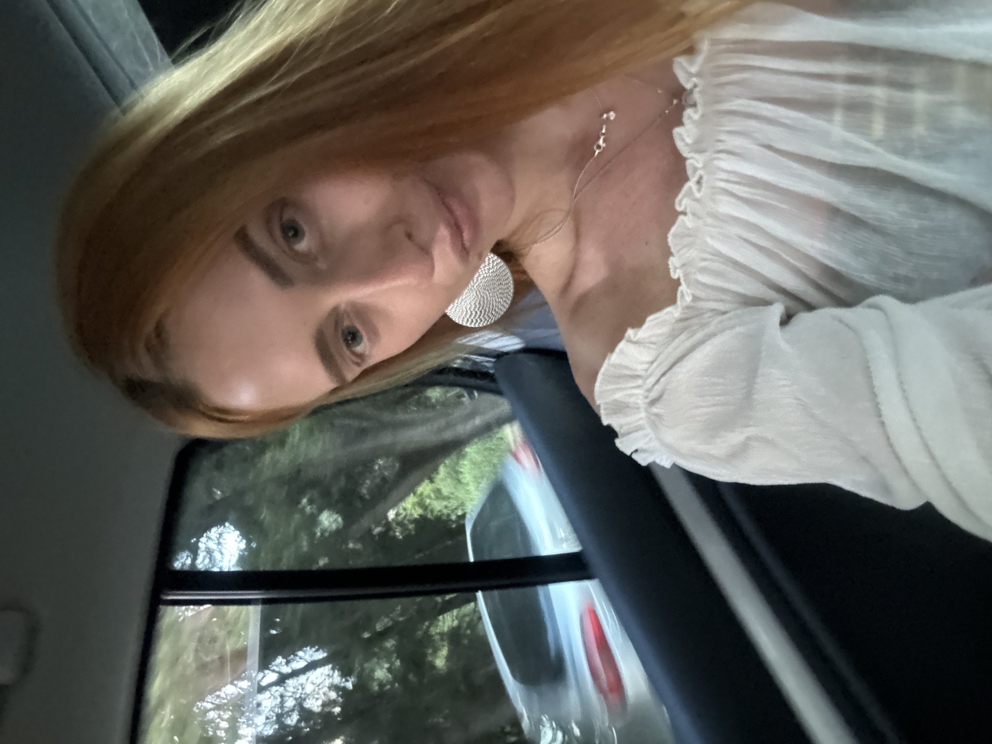 An upward-angled full-frame selfie captures a young adult woman with long, light brown hair and blue-grey eyes, seated in the passenger compartment of a vehicle in Petrovac centar, Montenegro. She gazes towards the camera with a subtle pout, her head slightly tilted. She wears a white, off-the-shoulder, ruffled top and a large, textured circular silver earring on her right ear, complemented by a delicate silver necklace with small charms. The car's dark interior trim and a window frame are visible to her left and behind her. Through the vehicle's window, motion-blurred green trees suggest a treelined road or parkway. A light-colored sedan is partially visible and motion-blurred in the background, indicating movement. The lighting suggests late afternoon or early evening.