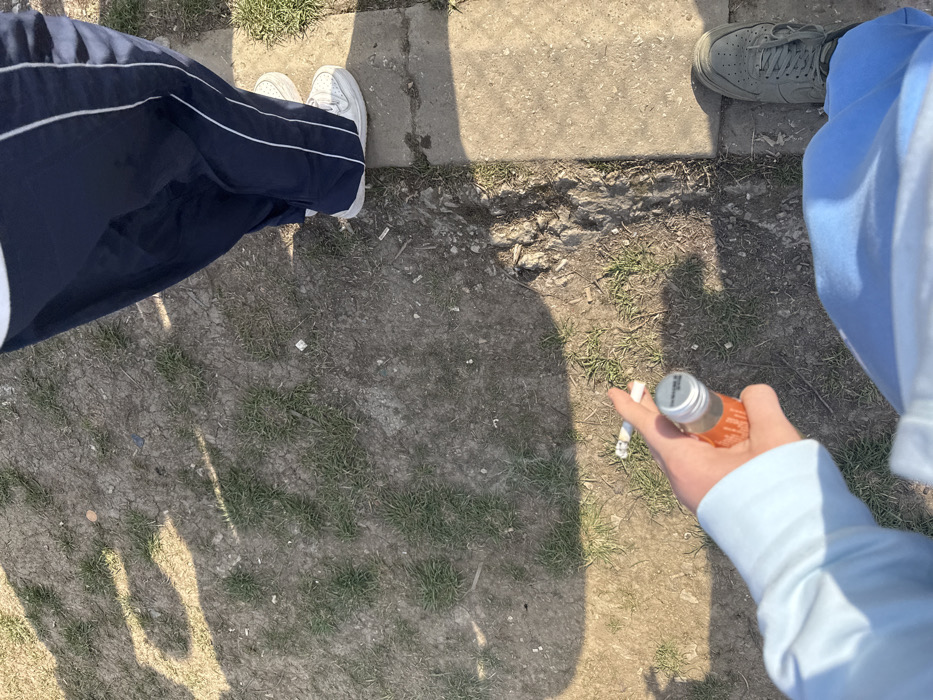 The image captures a ground-level scene in Troyan, Bulgaria, under bright daylight, with distinct shadows cast on the earth. The view is from a high angle, looking down.
On the right side of the frame, the lower half of an individual is visible, standing on a light grey concrete surface. This person is wearing dark blue track pants with a thin white stripe running vertically down each side and white athletic shoes, specifically white Nike Air Force 1s.
On the bottom left quadrant, the right hand and forearm of a second individual are visible. This person is wearing a light blue long-sleeved garment and light blue pants. The right hand holds two objects: a small, clear glass bottle containing an orange liquid, sealed with a silver-colored cap, and a partially consumed cigarette with an ash tip. The left foot of this second individual, clad in a dark grey or olive-green athletic shoe, is visible on the bottom right corner, positioned on both the concrete surface and the adjacent dirt.
The ground area, primarily in the center and left of the image, consists of dry, light brown soil with sparse patches of green grass. Scattered across this dirt are several discarded white cigarette butts and small pieces of litter. A distinct concrete curb or slab runs vertically along the right side of the frame, acting as a boundary between the dirt ground and the concrete surface.