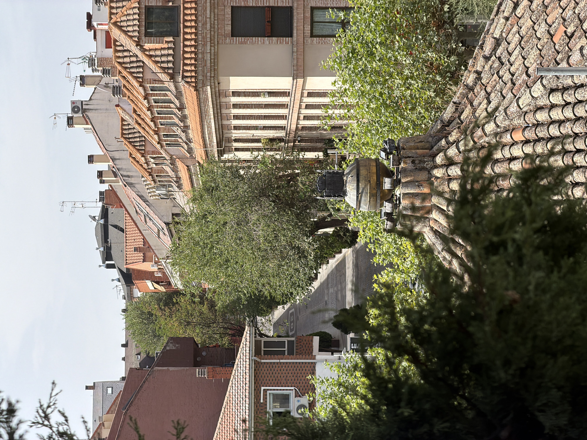 The image depicts a view overlooking a narrow, sloped street lined with trees and residential buildings in Las Rozas de Madrid, Spain. In the foreground, a tiled roof with a distinctive rounded chimney structure, adorned with multiple spotlights, is visible. The street itself has painted markings, including a "20" indicating a speed limit. On the left side of the street, a red brick house with white trim and windows is present. Across the street, multiple buildings with red tile roofs and a light-colored facade are seen, some featuring multiple stories and visible windows. Green trees with dense foliage flank the street and are interspersed among the buildings, casting shadows. The sky is overcast with a bright, diffused light. No people are visible in the frame.