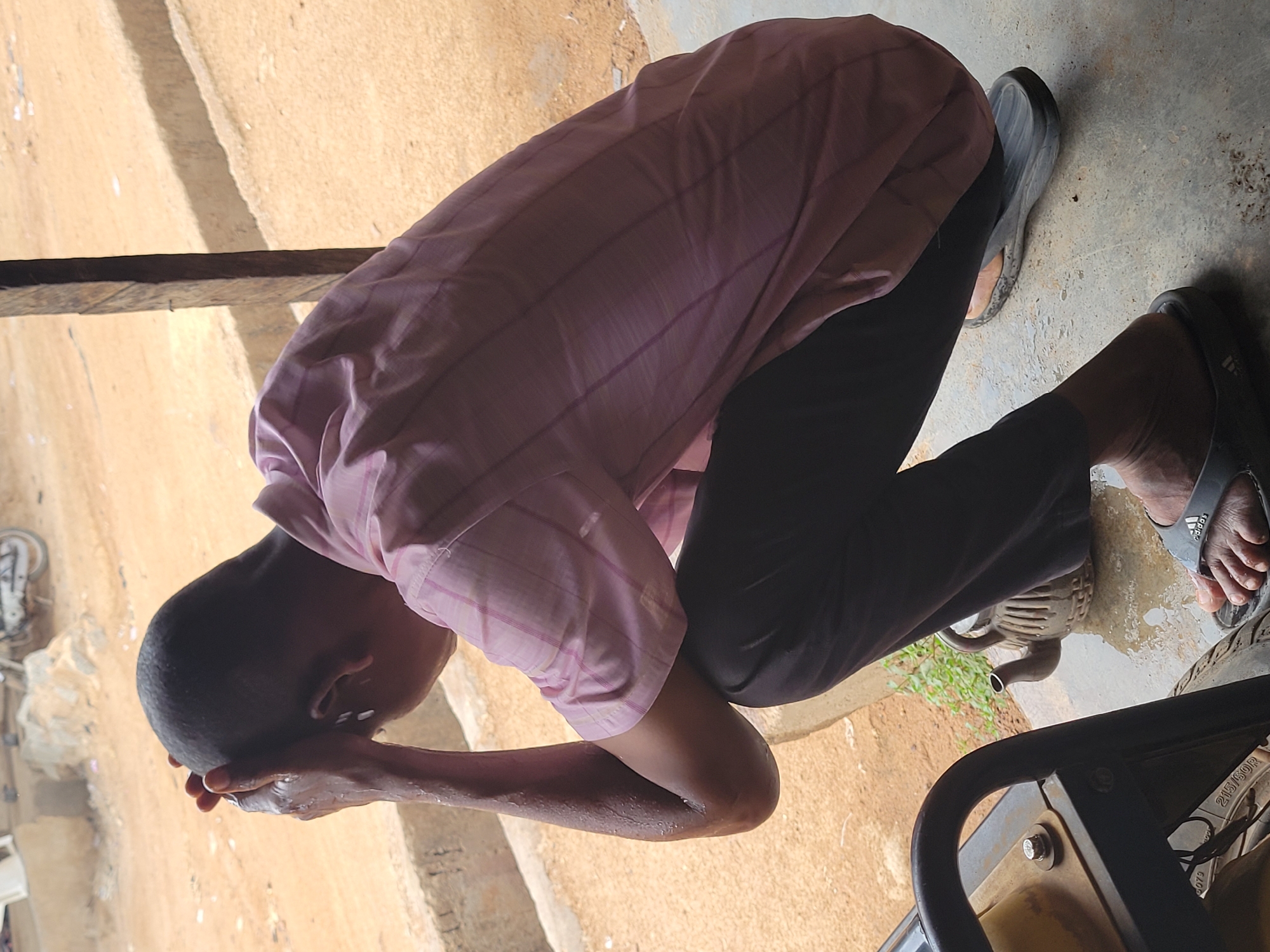 A person is washing their face outdoors in what appears to be Madalla, Nigeria. The individual is bent over, with their hands cupped to their face, presumably holding water. They are wearing a pink and white checkered short-sleeved shirt and black pants. They are wearing flip-flops on their feet. The ground is a mixture of concrete and dirt. A generator is visible in the lower right corner of the frame. A bicycle wheel is slightly visible in the upper left.