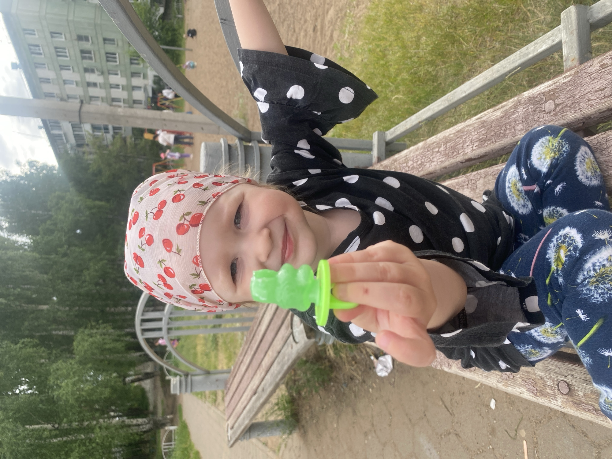A young girl wearing a pink headscarf with a cherry pattern and a black and white polka dot shirt is sitting on a wooden bench in a playground. She is holding a green plastic toy ring with a bear-shaped lollipop on it. Her pants have a dandelion print. In the background, there are trees, a playground with sand, and an apartment building. The photo was taken on a cloudy day.