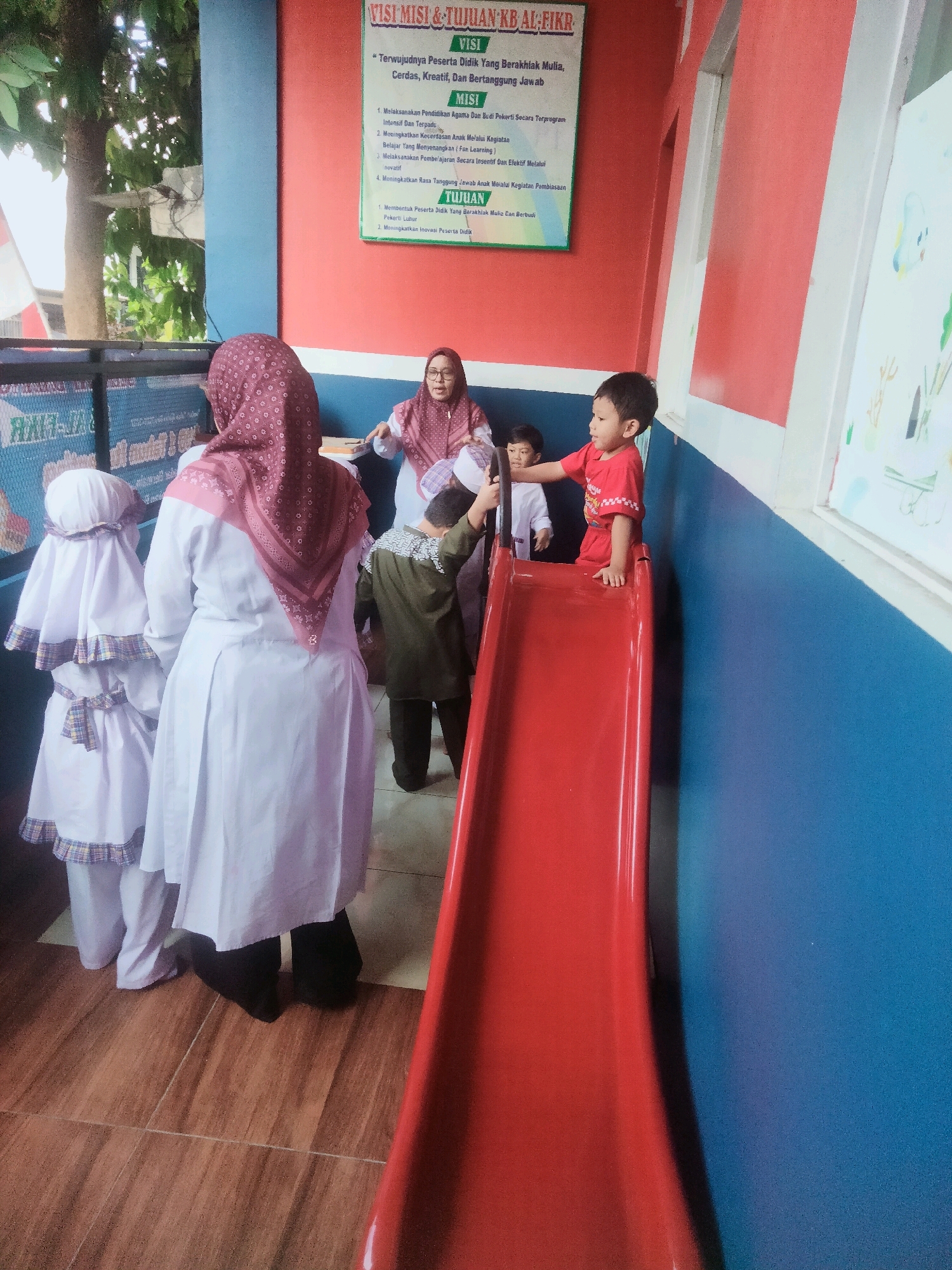 A group of individuals, including children, are gathered indoors near a bright red slide. Two children are in the foreground; one is wearing a red shirt and appears to be on the slide. Another child, wearing a white hijab and a long white garment, is standing behind the child on the slide. Further in the background, a woman in a pink hijab and glasses is interacting with a child in a green shirt and dark pants. Two other children in white garments and head coverings are to the left of the frame. The environment suggests a school or daycare setting, with a blue and red painted wall visible. A poster with Indonesian text is mounted on the red wall. The floor is tiled with a wood-like pattern. The scene appears to capture a moment of play or activity in a community space.