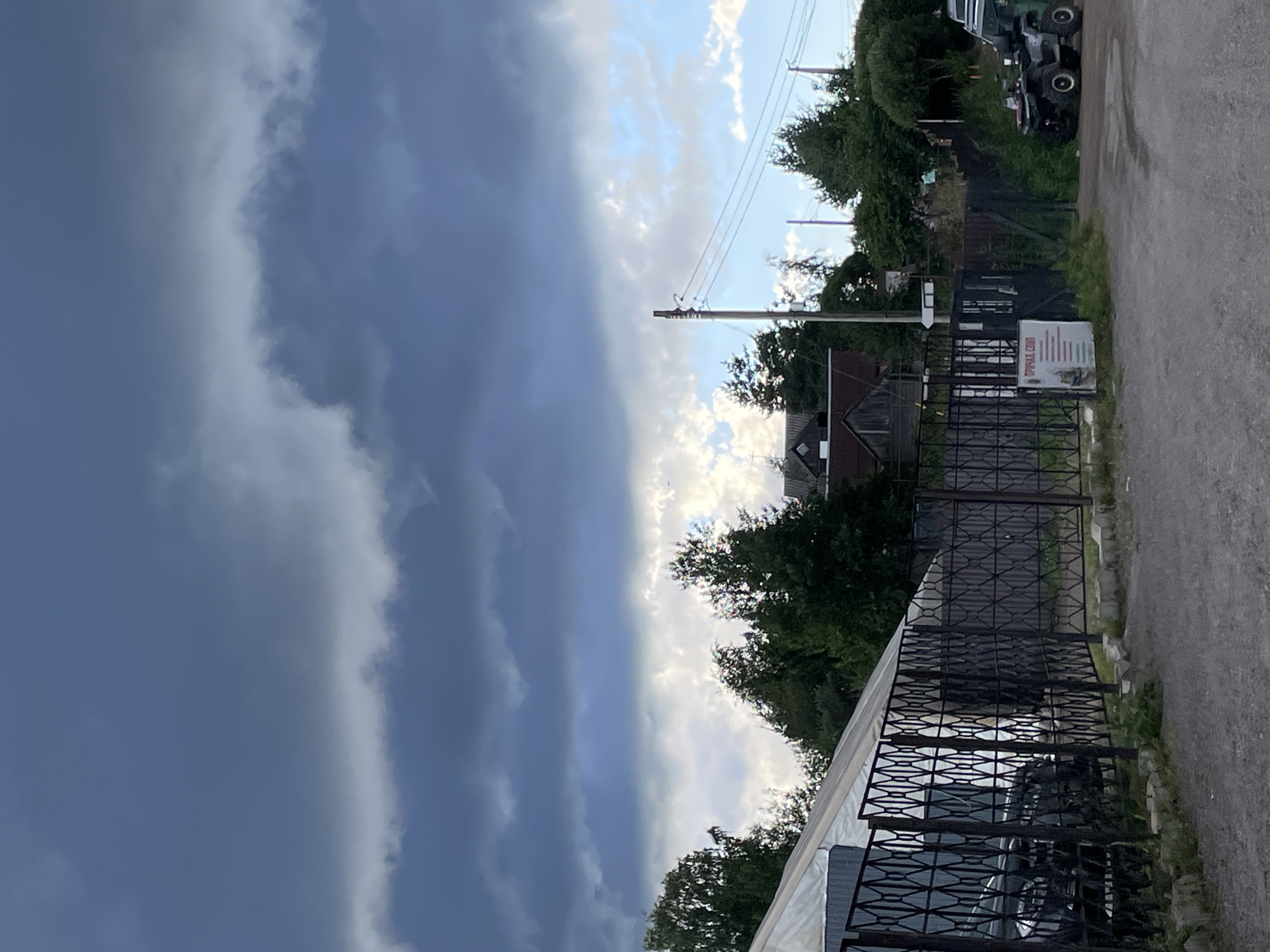The image depicts an outdoor scene in Berozovo, Russia, featuring a dark, cloudy sky overhead. In the foreground, a gravel area leads to a black metal fence with a diamond-shaped pattern. Beyond the fence, a large white structure, possibly a tent or temporary building, is visible. Adjacent to this structure, several dark-colored vehicles, including what appears to be a car and ATVs, are parked. Trees and some traditional Russian houses with dark roofs are scattered throughout the background. Power lines are visible stretching across the frame, supported by wooden poles. A white sign with red and green text is attached to the fence on the left side of the image. The overall impression is that of a somewhat overcast day in a semi-rural or industrial area.