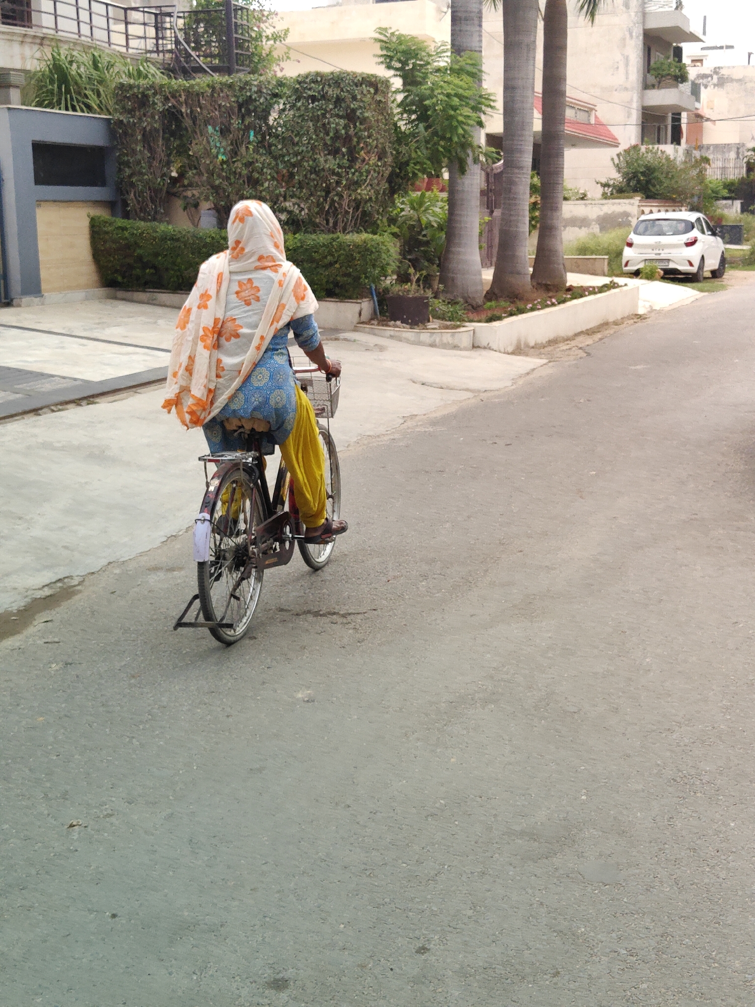 A woman wearing a blue salwar kameez and a floral white and orange dupatta is riding a bicycle. The woman is seen from the back, and she is cycling on an asphalt road. The bicycle has a basket at the front and a kickstand at the rear. The environment appears to be a residential area, with houses and manicured greenery visible in the background. A white car is parked on the right side of the road, and tall trees are present in the middle ground. The scene is likely in Gurgaon, India, given the architectural style and vegetation.