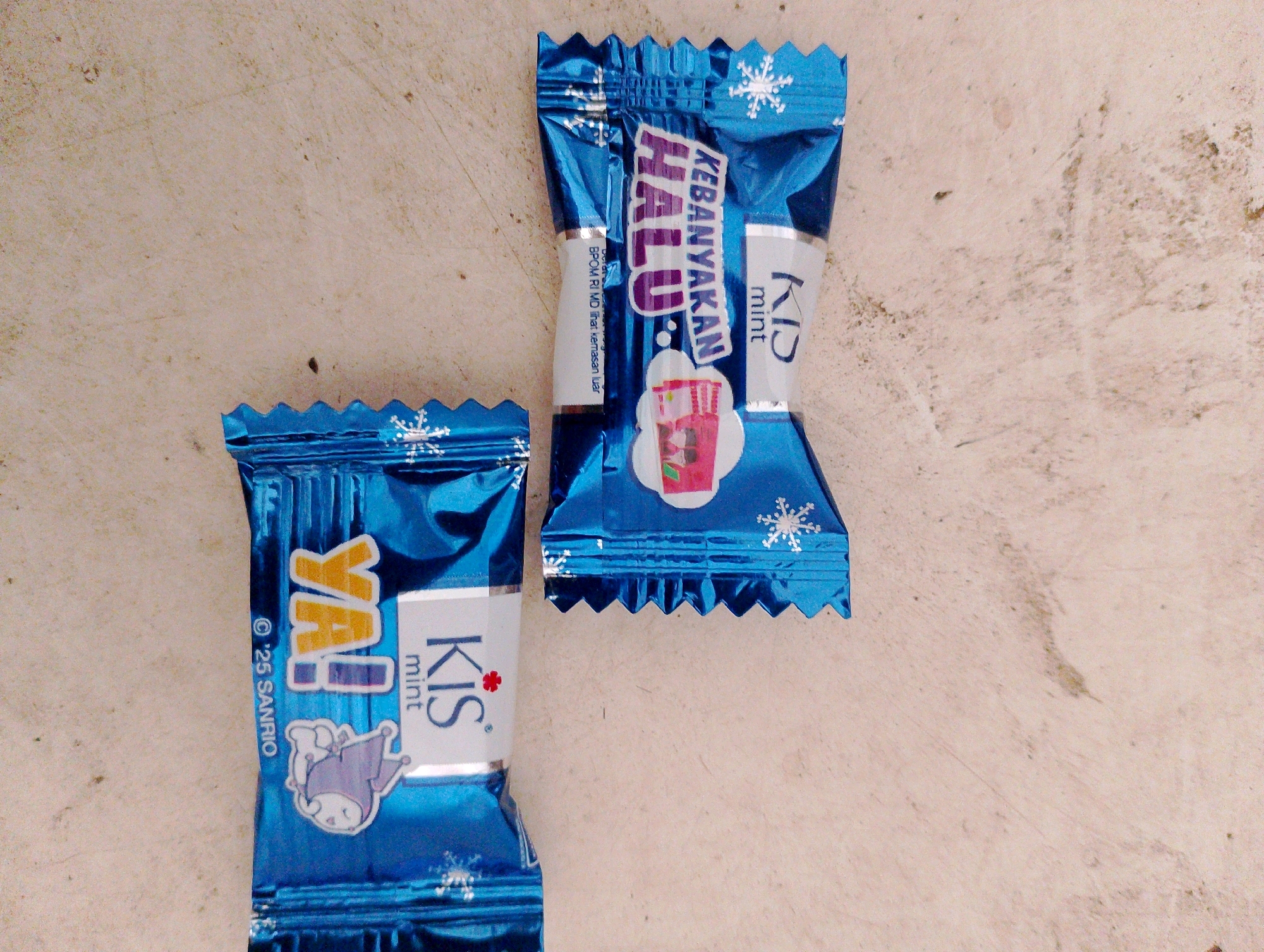 Two blue candy wrappers, one labeled "HALU" and the other "YAO!", lie on a textured, light-colored surface. The "HALU" wrapper, on the right, features the text "KEBANYAKAN HALU" in purple and white lettering, with "KID" and "mint" also visible. A depiction of a pink drink with a straw is on this wrapper, along with snowflakes. The "YAO!" wrapper, on the left, has "YAO!" in yellow and white lettering, and the words "Kis" and "mint" are also present. A drawing of a character resembling Hello Kitty's friend, My Melody, is on this wrapper, along with snowflakes and the text "'25 SANRIO". The background surface appears to be a concrete or stone surface, with some dust and discoloration. No specific location in Merauke, Indonesia, can be determined from the image.