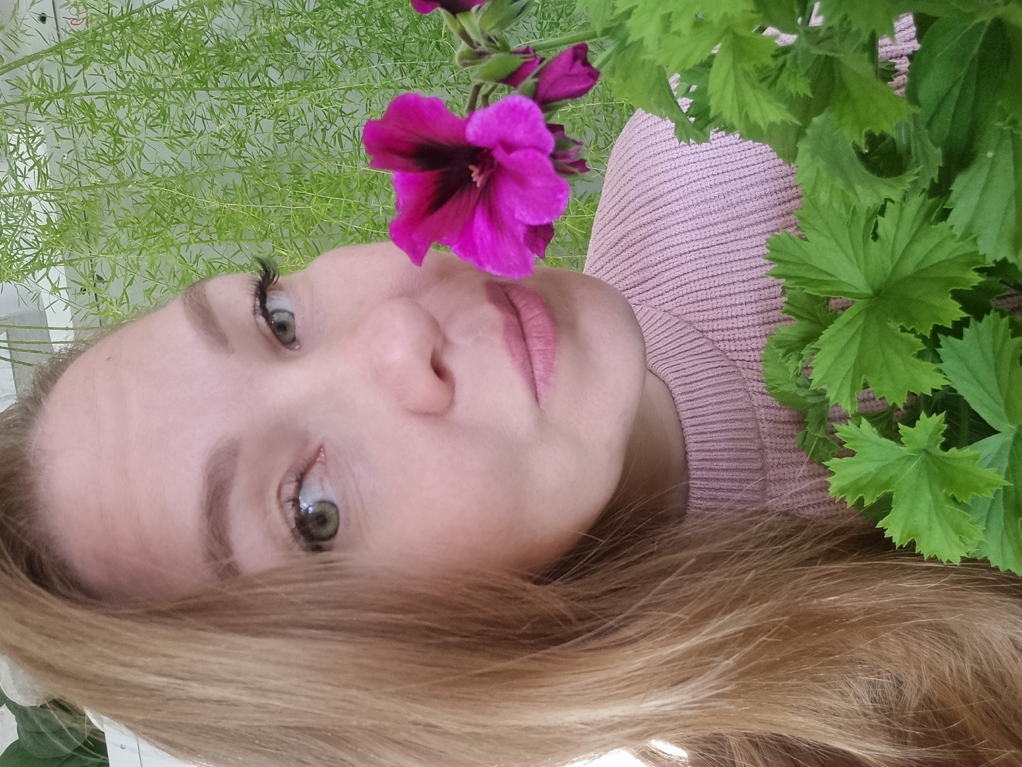 The image shows a woman's face in close-up, turned at an angle. Her complexion is fair, and she has light-colored hair. A bright pink flower is positioned near her face. Green foliage surrounds her. She wears a light pink sweater. The background includes delicate, fern-like greenery. The location of the photo is not provided.