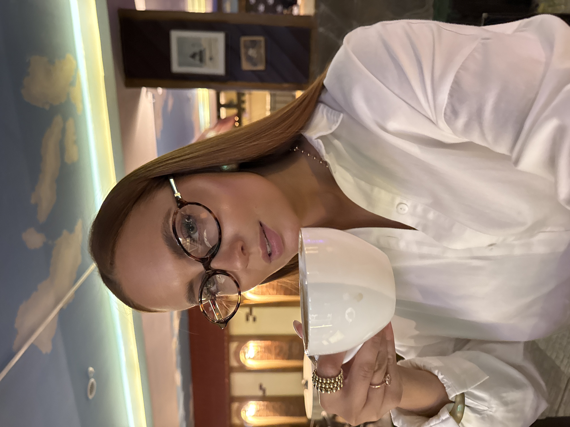 A woman with auburn hair and glasses sits inside what appears to be a café or restaurant, holding a white cup. She is wearing a white, buttoned-down shirt and has several rings on her fingers. The background suggests an interior setting with framed pictures on the wall and a uniquely decorated ceiling that includes a depiction of clouds and sky. Given the request to specify location details in Chaikovsky, Russia, if available, the image might have been taken in such a cafe or restaurant. However, without more specific location cues or a clear indication of the city, confirming it to be Chaikovsky, Russia is not possible.