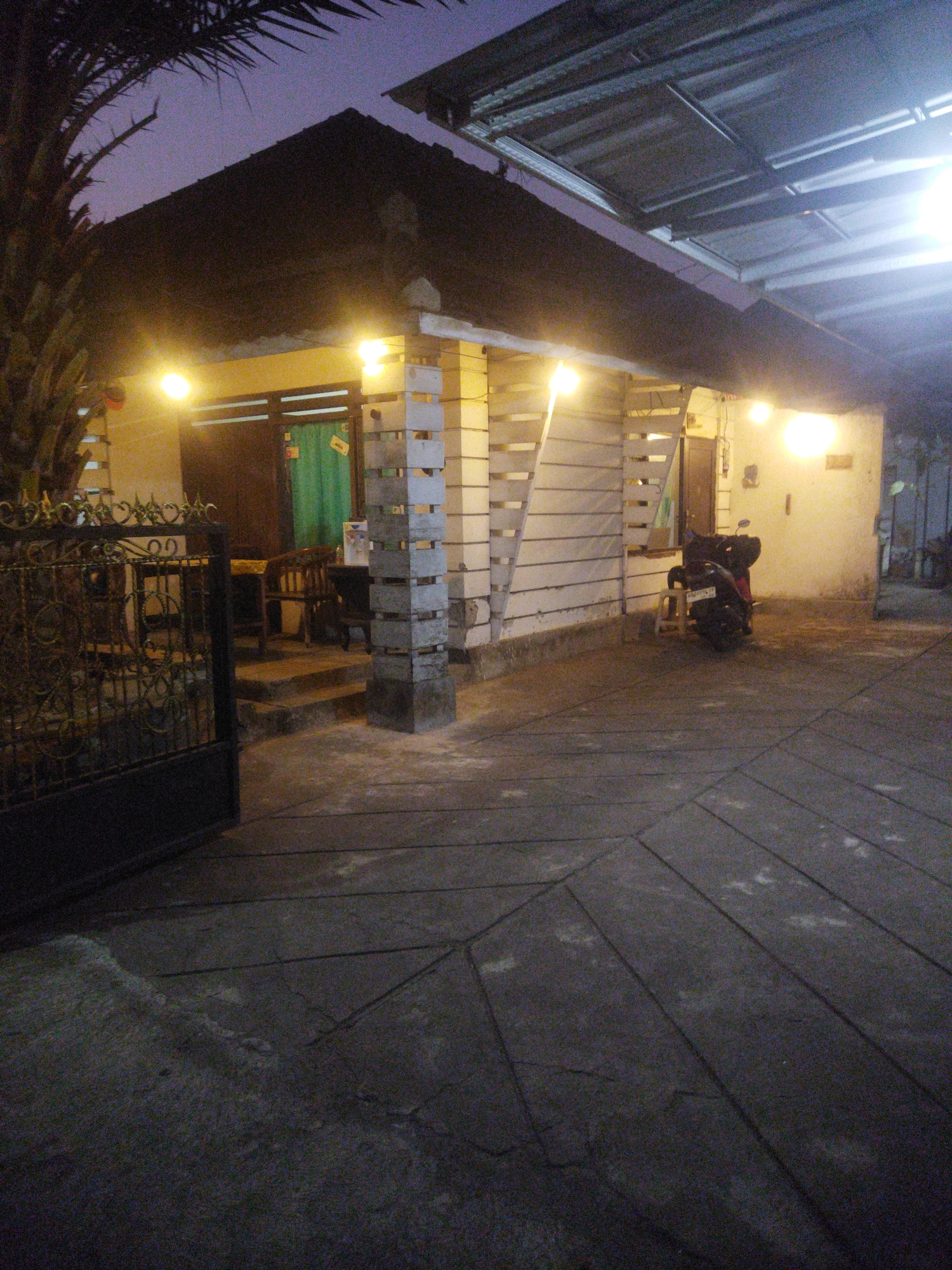 The image depicts a dimly lit scene outside a residential building at dusk or night in Wonogiri, Indonesia. A dark-colored motorcycle with a red seat is parked on the paved ground in front of a light-colored wall. To the left, a black wrought-iron gate stands partially open, revealing a glimpse of a covered seating area with a table and chairs. The building features a dark-colored roof and is illuminated by several warm-toned lights. A palm tree is visible on the far left of the frame. The ground is paved with large, irregularly shaped concrete slabs. The overall atmosphere is quiet and residential.