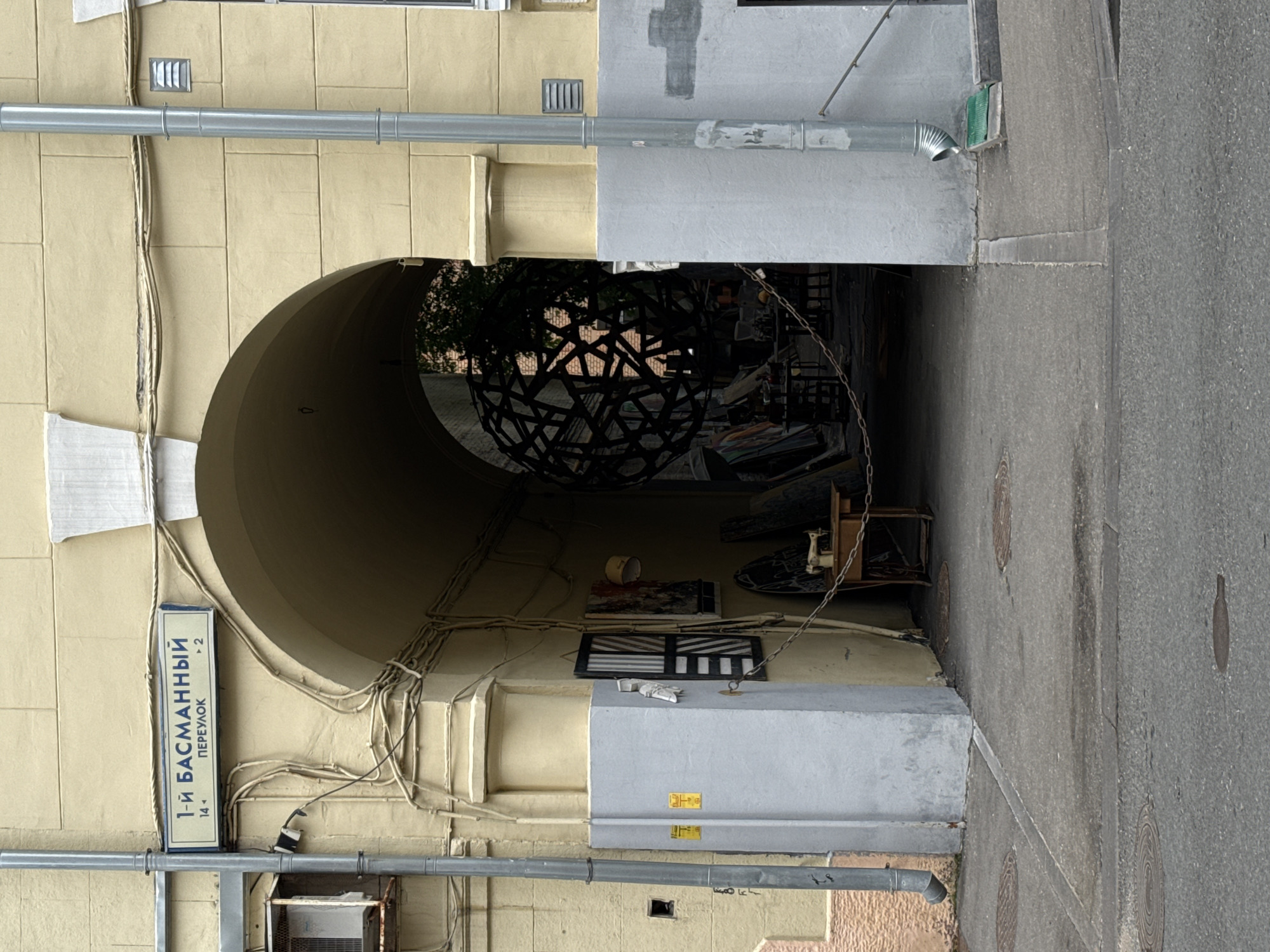 The image depicts an archway leading into a courtyard in Moscow, Russia. A street sign above the archway reads "1-Й БАСМАННЫЙ ПЕРЕУЛОК" (1st Basmanny Lane), with numbers "14" and "2" indicating building numbers or range. The archway is constructed from light yellow masonry, with white decorative elements at the top. Two grey downspouts are visible on the exterior walls.
Inside the courtyard, a large, dark, spherical metal sculpture is hanging. Beneath it, a variety of objects are visible, including what appears to be an old sewing machine on a wooden table, several canvases or artworks leaning against a wall, and other assorted items suggesting a workshop or storage area. A chain is draped across the courtyard. The ground inside the courtyard appears to be concrete, with some circular manhole covers visible on the pavement leading up to the archway. The overall impression is of an urban space with artistic or industrial elements.