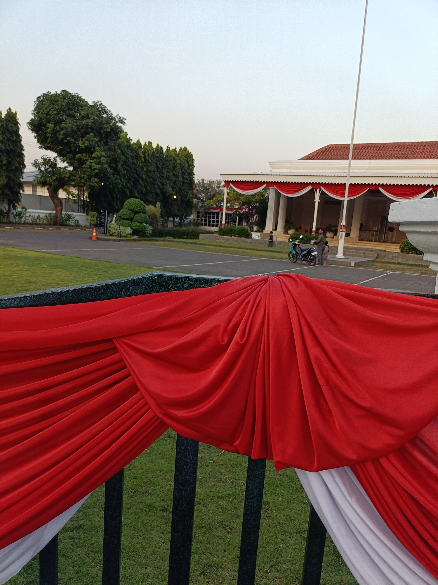 The image shows a view of the front of the Gedung Gubernuran, the governor's office in Semarang, Indonesia. The building is decorated with red and white draped fabric, which is also visible in the foreground of the image. A flagpole stands in front of the building, and a motorcycle with two people on it is parked nearby. The sky is clear and overcast. There is a well-maintained lawn in front of the building, and trees and other greenery surround the area. The scene appears to be set during the daytime.