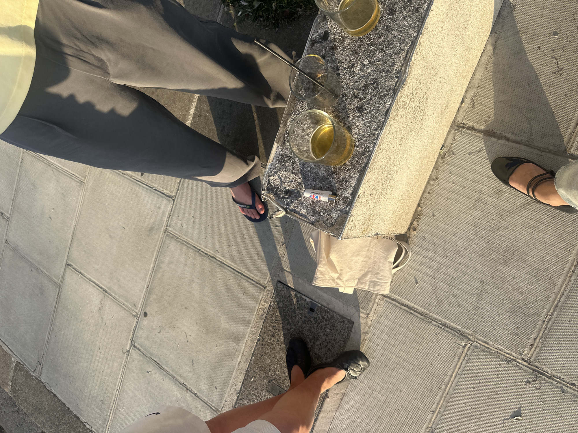 The scene captures a casual gathering in what appears to be a public space. Three individuals are partially visible. One person is wearing gray trousers and blue flip-flops. Adjacent to them is a gray bench-like structure with three glasses containing yellow liquid, possibly beverages, and a metal straw. Next to these glasses is a small, rectangular item with red, white, and blue markings. A canvas tote bag is situated near the bench. A second person is wearing black shoes and a white garment. The third person is wearing brown sandals. The setting is a paved area with geometric tile patterns. The location is likely in London, United Kingdom, based on the style of the paving stones and the tote bag.