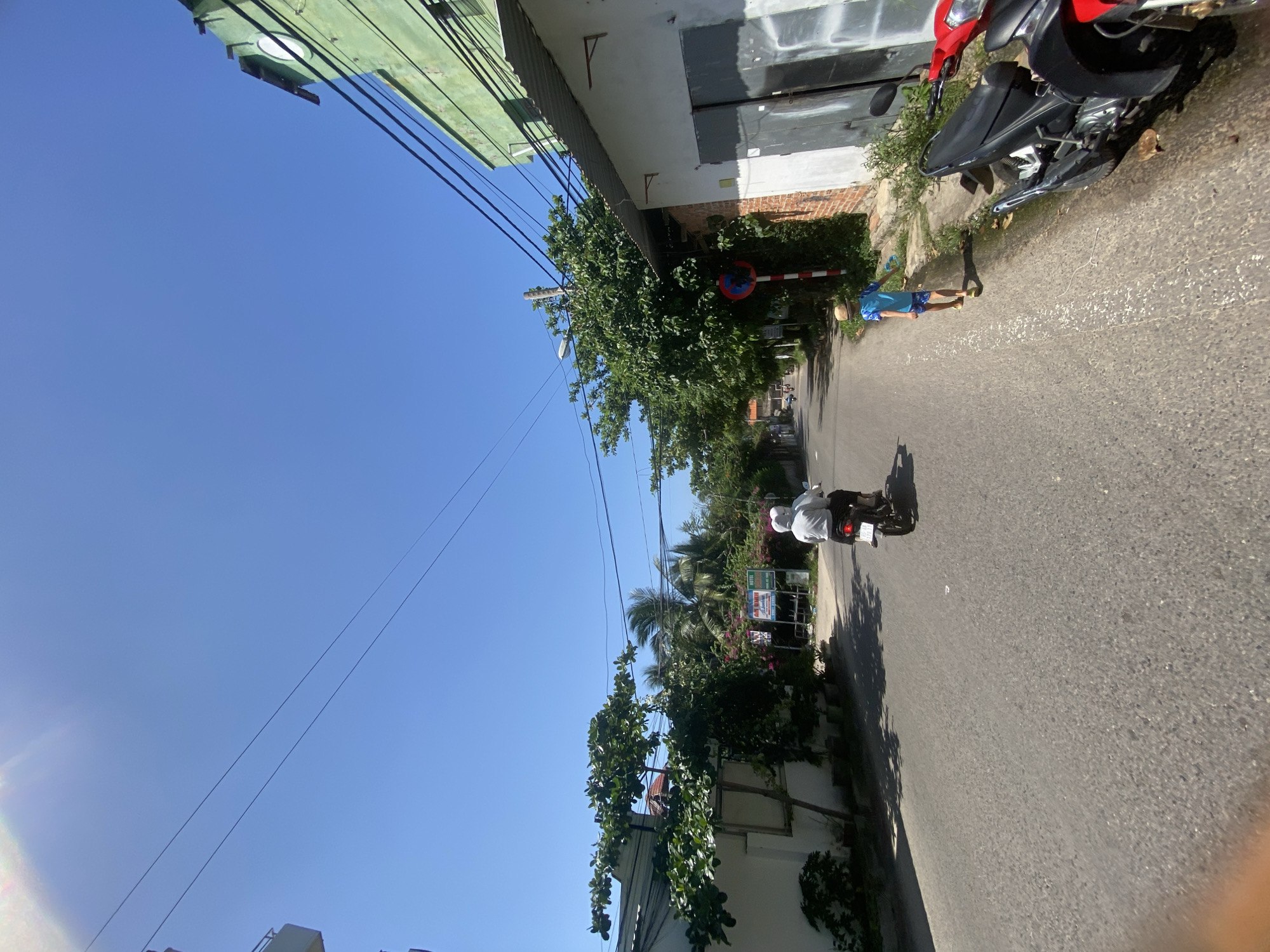 The image depicts a street scene in Mũi Né, Vietnam, under a clear blue sky. In the foreground, a gray asphalt road stretches into the distance, flanked by buildings and vegetation. On the right side of the road, a red and black motorcycle is parked. Further down the road, a child wearing a straw hat and blue and white patterned shorts is walking away from the camera. On the left side of the road, a person on a scooter, wearing a white head covering and a light-colored top, is riding away from the camera. Behind the scooter rider, a building with a white exterior and some greenery is visible. Trees and tropical foliage line the sides of the street, with power lines crisscrossing overhead. To the right, a green building with a corrugated metal roof and a white wall with large gray doors is prominent. A red and white striped traffic sign is mounted on a pole near the green building. Signs advertising "AN NHI" and "HOTEL" are visible in the distance on the left side of the road, suggesting a commercial area. Shadows from the trees are cast across the road.