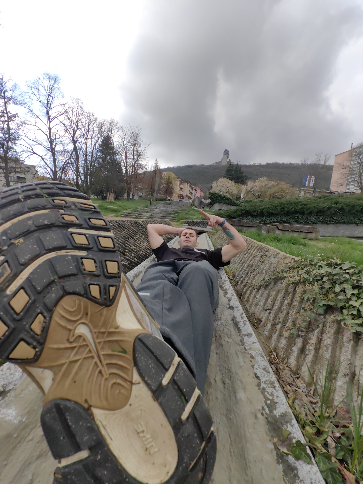 A low-angle, close-up shot captures a person lying in a concrete channel within the city of Shumen, Bulgaria. The person is lying on their back, wearing a black shirt and gray pants, with hands behind their head and one arm raised pointing toward the top right of the frame. A prominent tattoo is visible on their forearm. A large Nike shoe fills the lower left corner, its sole facing the camera, appearing worn and dirty.
In the background, beyond the concrete channel, is a park-like setting with trees, grass, and some buildings. A monument is visible on a distant hilltop, partially obscured by a cloudy, overcast sky. The overall perspective is distorted due to the wide-angle lens. The scene suggests an outdoor setting, likely a public space, with a combination of natural elements and urban infrastructure.