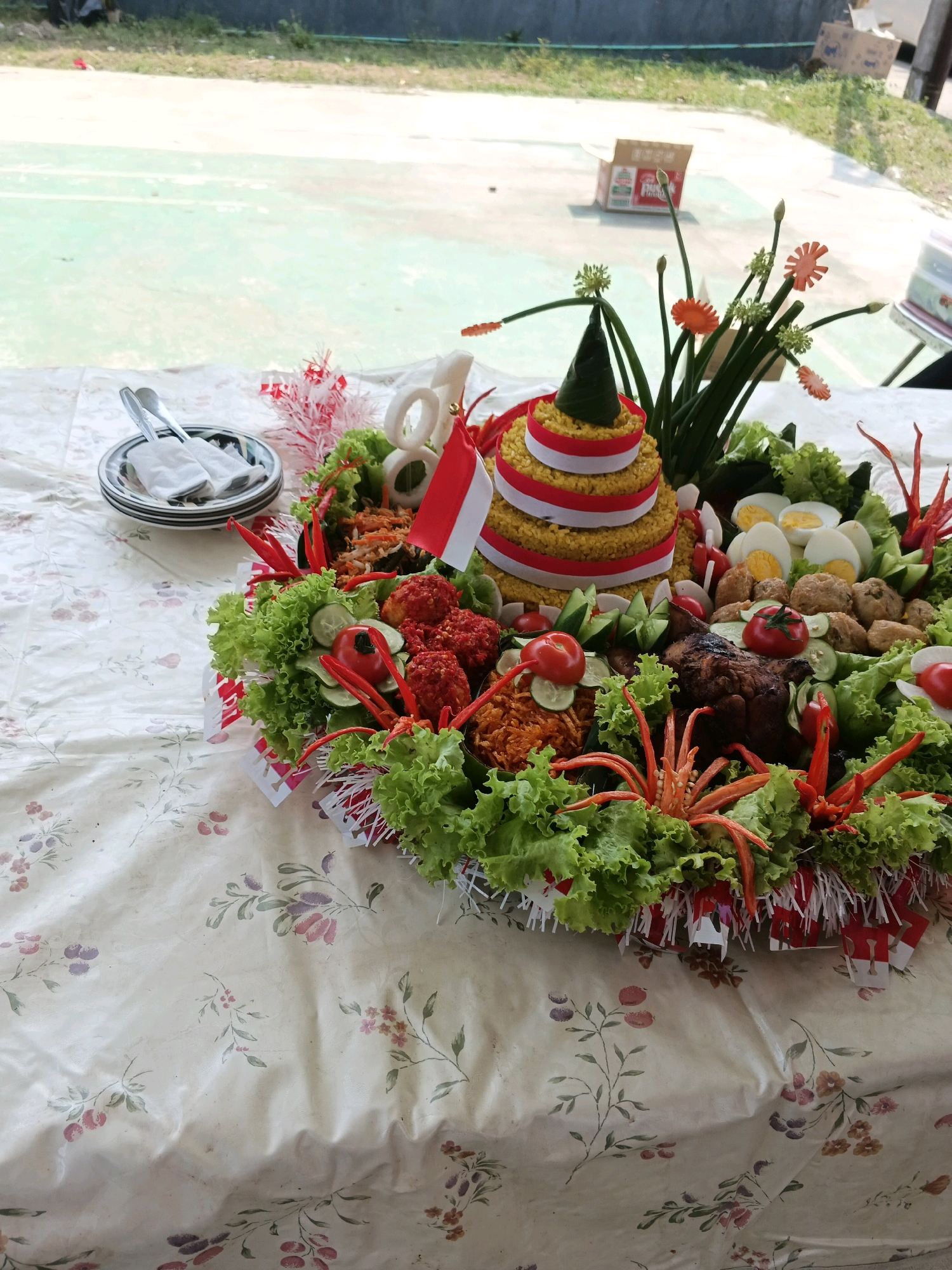The image depicts a food arrangement, likely a Nasi Tumpeng, displayed on a table covered with a floral-patterned tablecloth. The Nasi Tumpeng is conical in shape and decorated with red and white ribbons, resembling the Indonesian flag. It is surrounded by various side dishes, including fried chicken, hard-boiled eggs, vegetables like lettuce and cucumbers, and red chili peppers. Decorative elements such as green onions and orange carrot flowers are also present. To the left of the Nasi Tumpeng, a stack of plates and cutlery is visible. The background suggests an outdoor setting, possibly a courtyard or open area, with a glimpse of a green surface, possibly a sports court, and some boxes in the distance. The lighting indicates it is daytime. The scene is devoid of people, focusing entirely on the food presentation. The location is identified as Margahayu, Indonesia, implying this is a celebratory or traditional Indonesian meal setting.