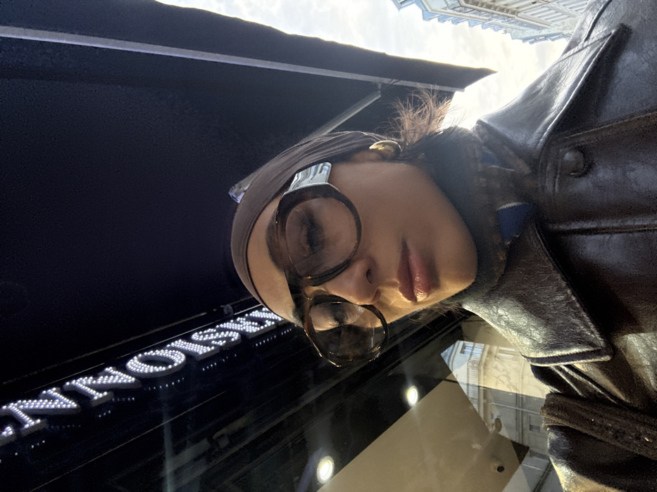 A woman in a brown leather jacket and headscarf poses for a selfie. Oversized glasses with a light brown frame dominate her face. The backdrop features an awning and the reflected facade of a building. A storefront sign with illuminated letters is also visible. The image appears to have been taken from a low angle, pointing upwards.