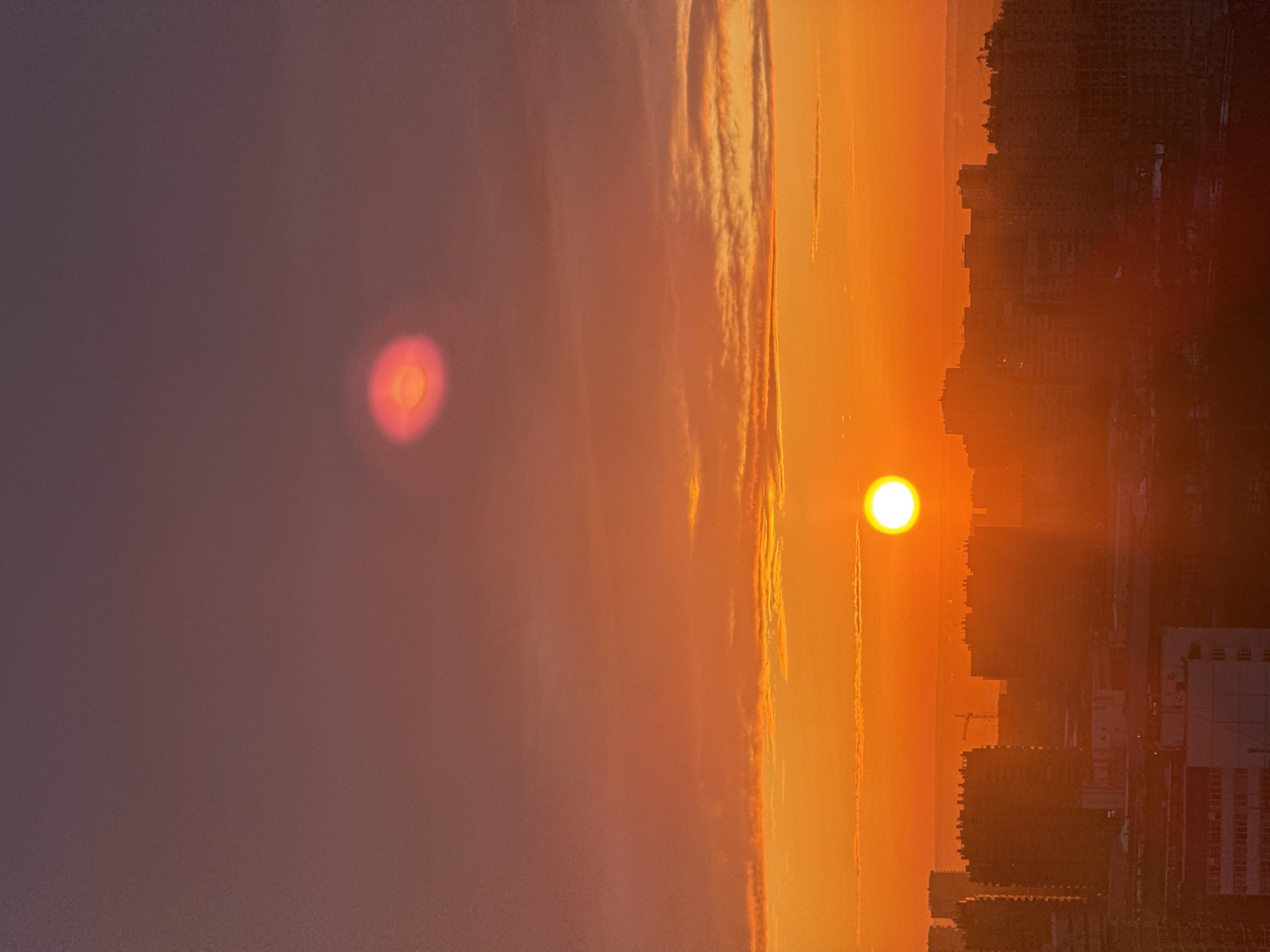 The image displays an elevated cityscape view of Perm, Russia, during either sunrise or sunset. The sky presents a color gradient from intense orange and red near the horizon to a deep purplish-mauve in the upper portion. A distinct, bright, circular sun, appearing white-yellow, is visible slightly above the urban skyline and to the left of the frame's vertical center. Above the sun, a prominent, diffuse, reddish-pink circular optical artifact or atmospheric light phenomenon, featuring a slightly darker, ring-like center, is present. The lower half of the frame is occupied by the dark silhouettes of numerous multi-story buildings, forming a dense urban horizon line. Faint structural details of buildings and at least one construction crane are discernible within this silhouetted mass. No human subjects, specific actions, or interactions are visible.
