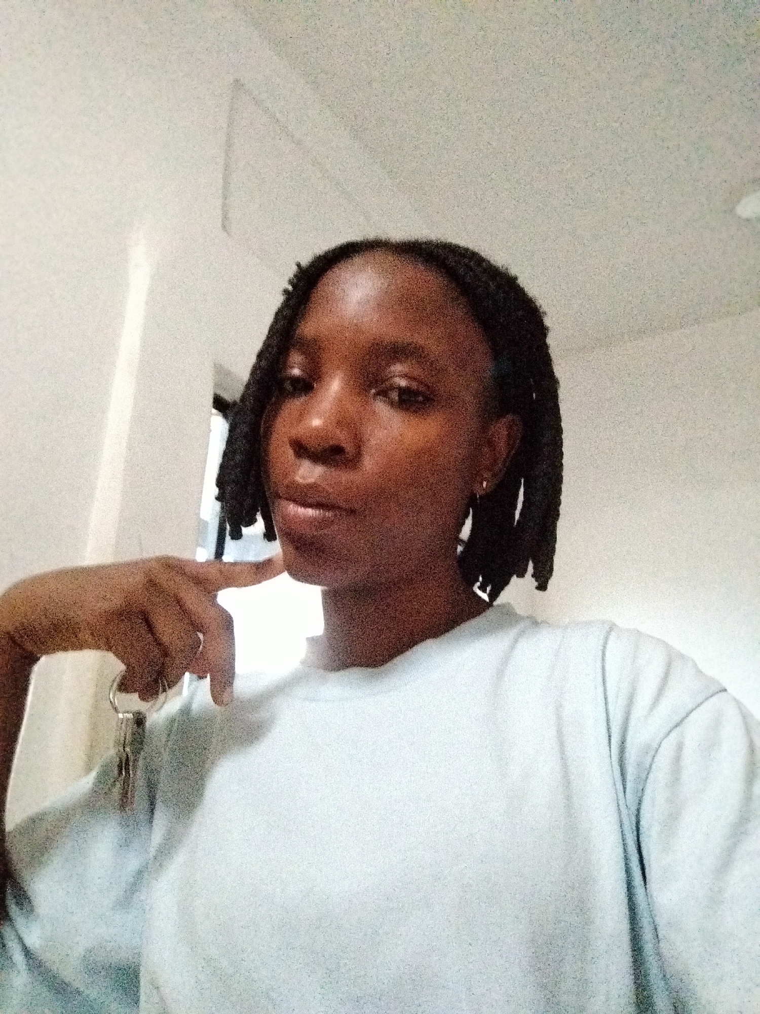 A young woman with braided hair and a light blue t-shirt is shown from the chest up, holding a set of keys and a ring on her left hand. Her right hand is positioned near her face, with her index finger touching her chin. The background appears to be an indoor setting, possibly a room in Uselu, Nigeria, with a white wall and a faint shadow. The lighting suggests it may be daytime, with light coming from the left. The image is a selfie.