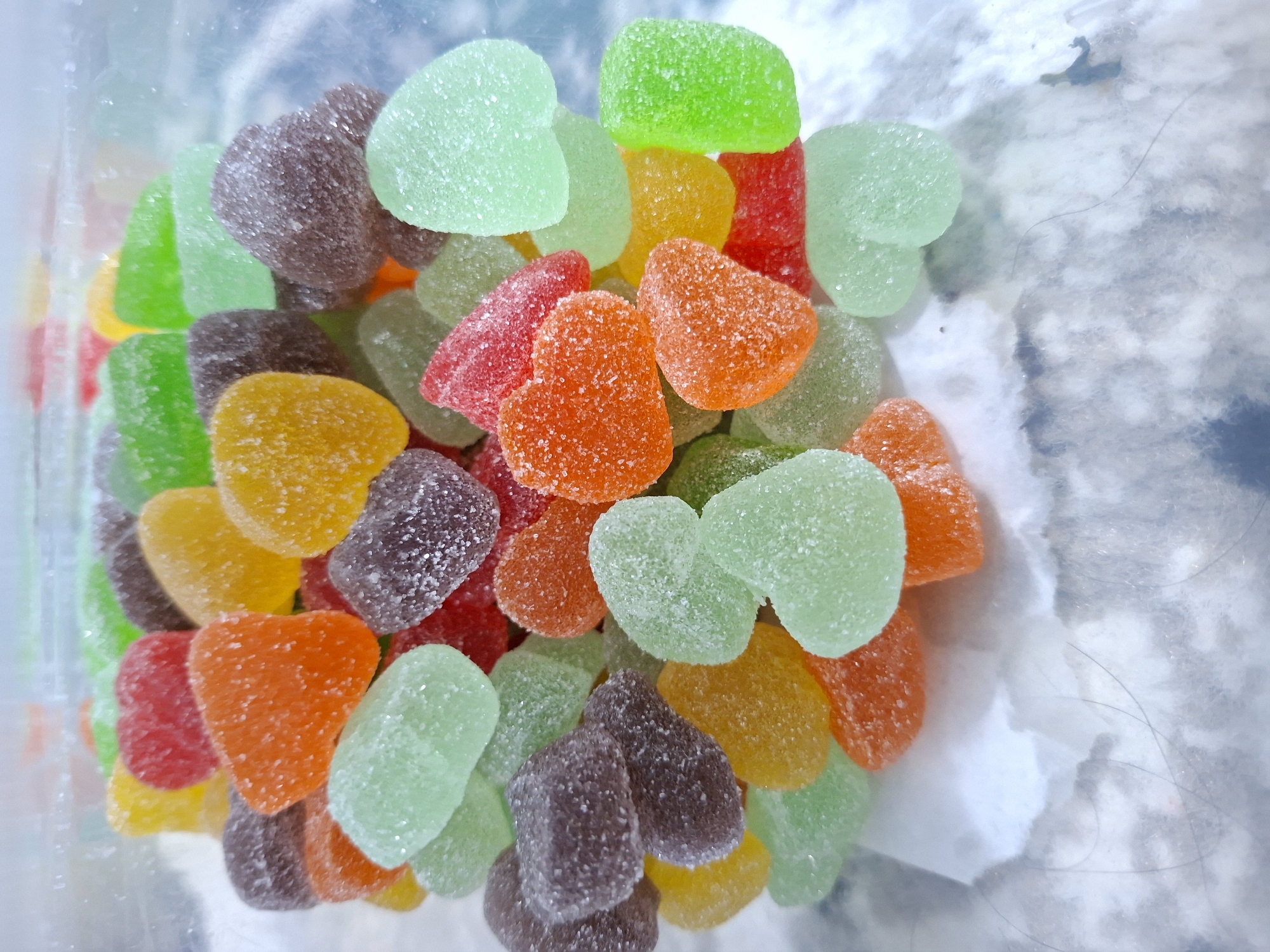 A close-up, overhead shot shows a pile of heart-shaped gummy candies in a clear plastic container. The candies are various colors, including green, orange, red, yellow, and purple. They have a granular, sugar-coated texture. The container appears to be partially empty, with a cloudy, white surface visible underneath the candies. A few stray hairs are also visible on the clear surface of the container. The location is Kutaglumpang, Indonesia.