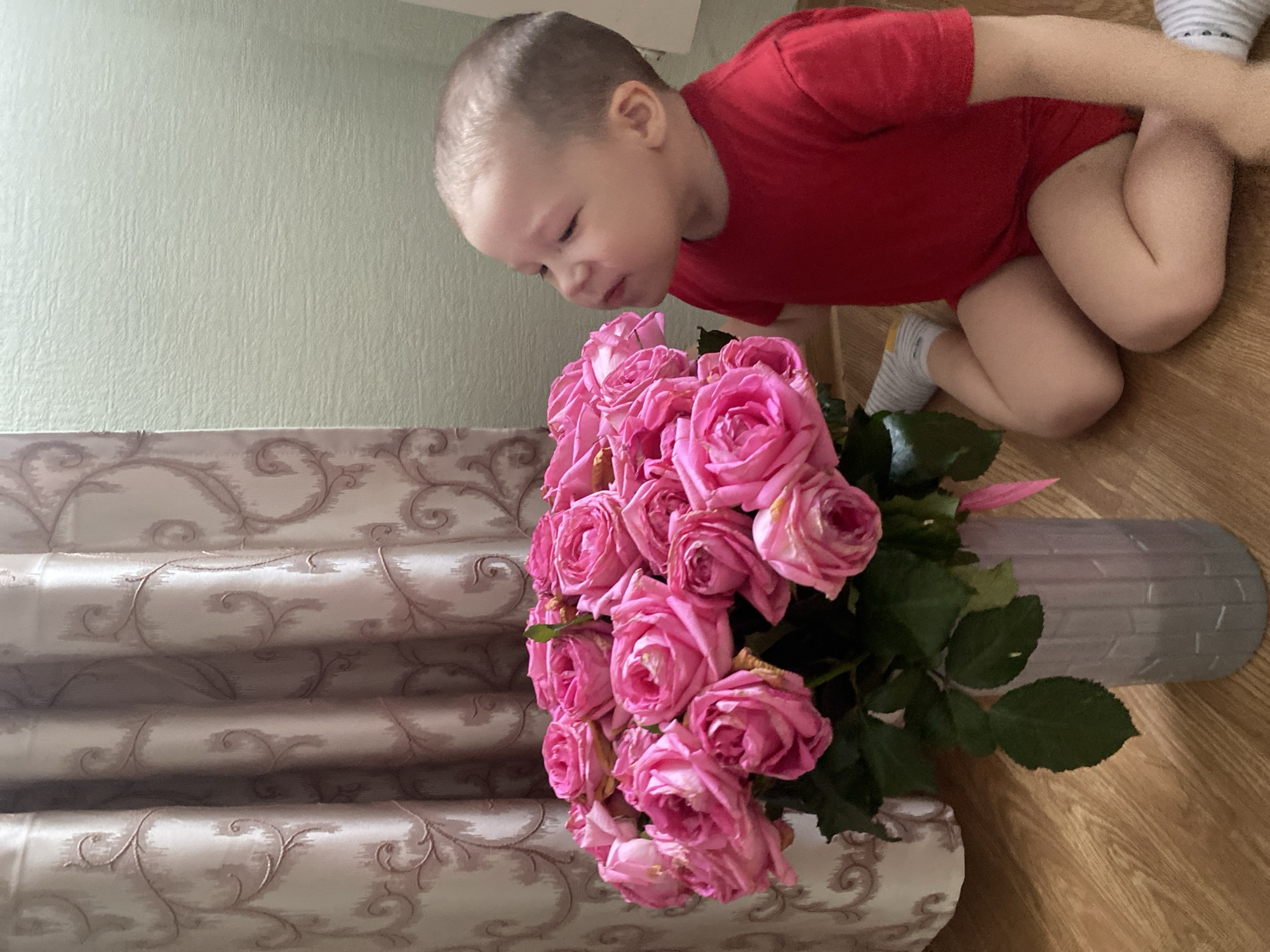 A child in a red t-shirt and shorts is bending over a bouquet of pink roses. The bouquet is in a light gray vase with a brick-like pattern. The child is on a wooden floor next to a patterned curtain. The scene is located in Sochi, Russia.