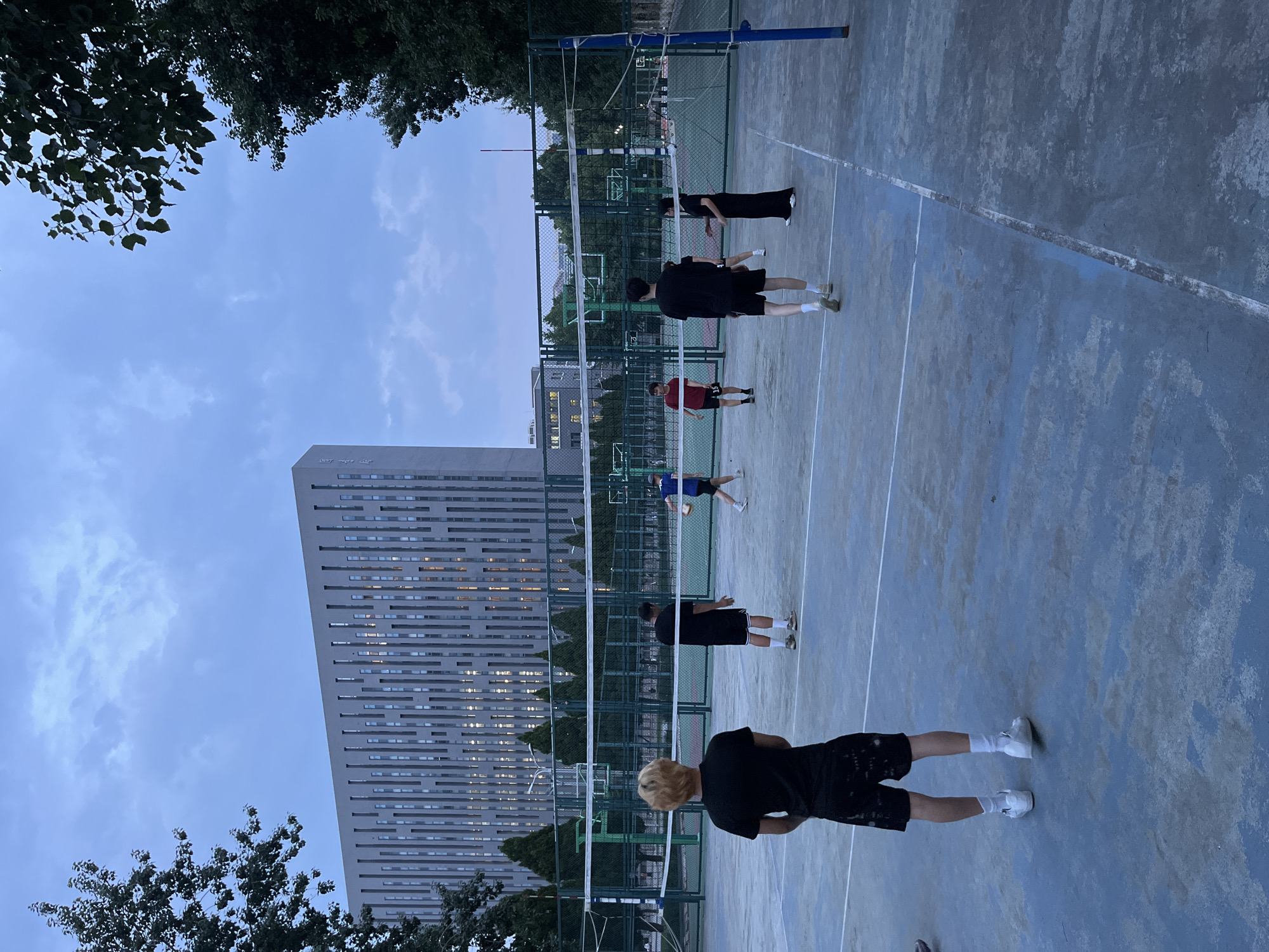 The image depicts an outdoor volleyball court with several young people engaged in a game or practice. The court is marked with white lines on a blue-grey surface. A net is set up across the middle of the court. The subjects are dressed in casual athletic wear, primarily black and dark colors. One person in the foreground, facing away from the camera, has blonde hair and is wearing a black t-shirt and patterned shorts. Other individuals are scattered across the court, some appearing to be in motion.
In the background, a tall, modern building with many horizontal windows is visible, suggesting an urban or campus setting. Trees frame the scene on the left and top edges. The sky is overcast, with hints of clouds, indicating it is likely late afternoon or early evening, shortly after sunset. The lighting suggests a dim, twilight atmosphere. The scene appears to be in Daxing District, China, given the provided location context. There is no visible text in the image. The overall mood seems relaxed and active, consistent with a sports activity among friends.